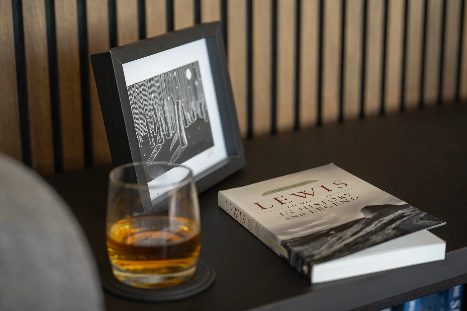 A close-up of a cozy living room scene featuring a book, a glass of whiskey, and a small picture frame on a TV stand. This inviting arrangement captures a sense of warmth and relaxation, perfect for unwinding with a good read and a drink in a comfort