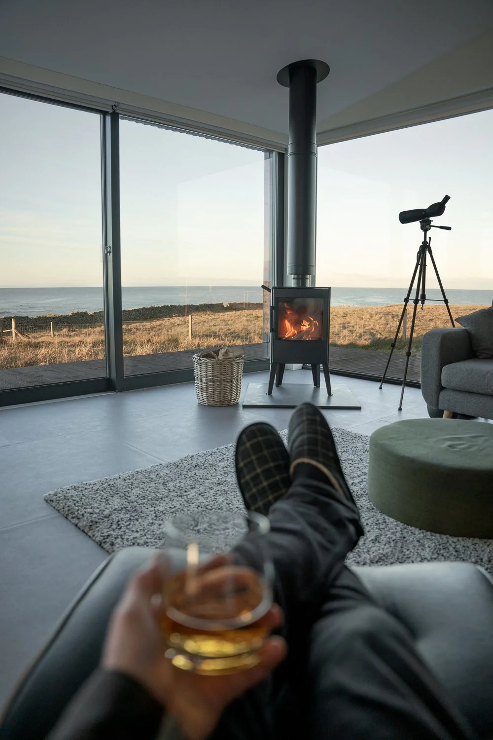 A cozy POV of a person sitting in an armchair, holding a glass of whiskey in front of a crackling fireplace. The warm glow of the fire contrasts beautifully with the stunning views visible through the floor-to-ceiling windows behind the fireplace. 