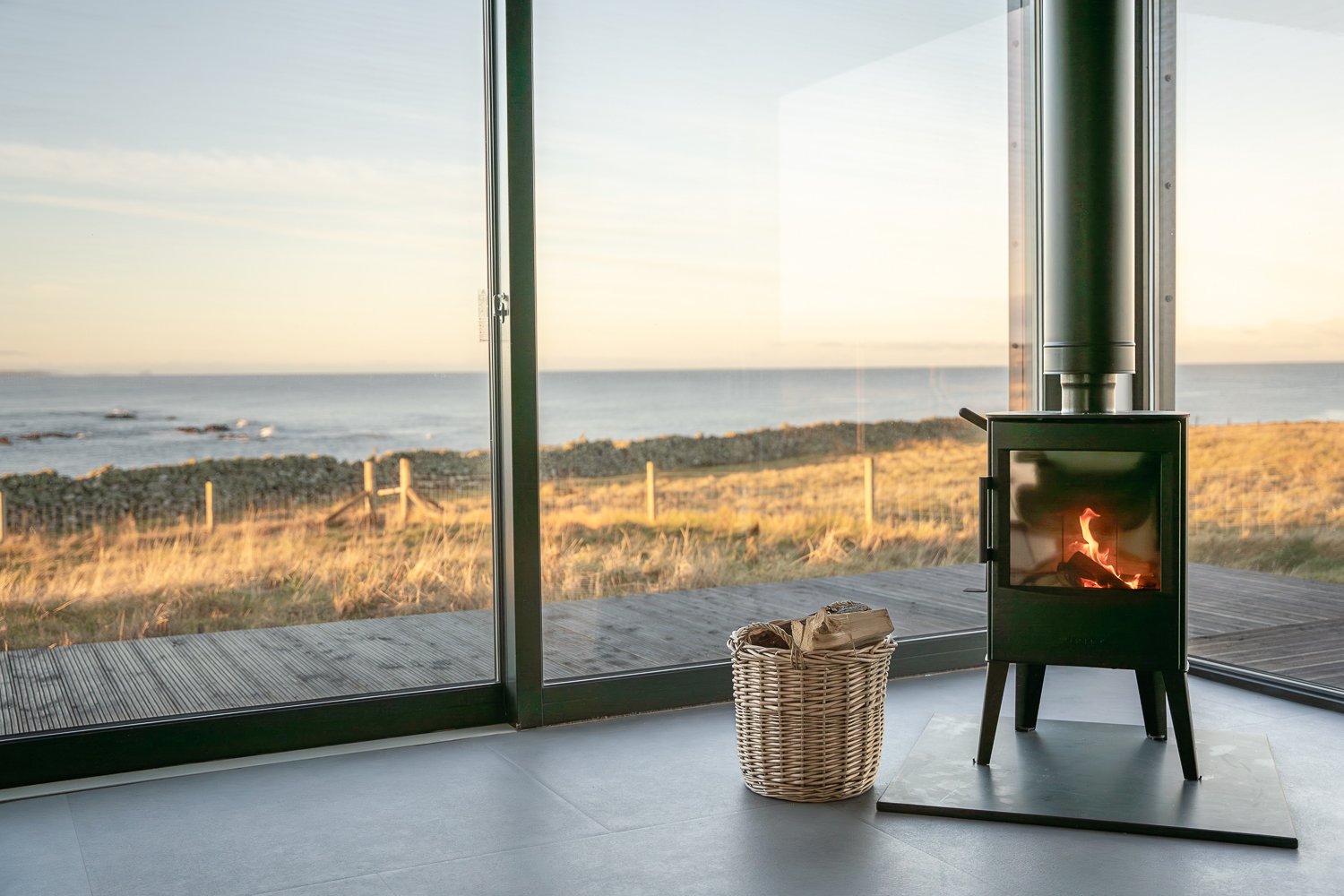 Cozy living room with a modern wood-burning stove by a large glass window overlooking a scenic coastal view at sunset on the Isle of Lewis, Scotland. The interior features a wicker basket filled with firewood, creating a warm and inviting atmosphere.