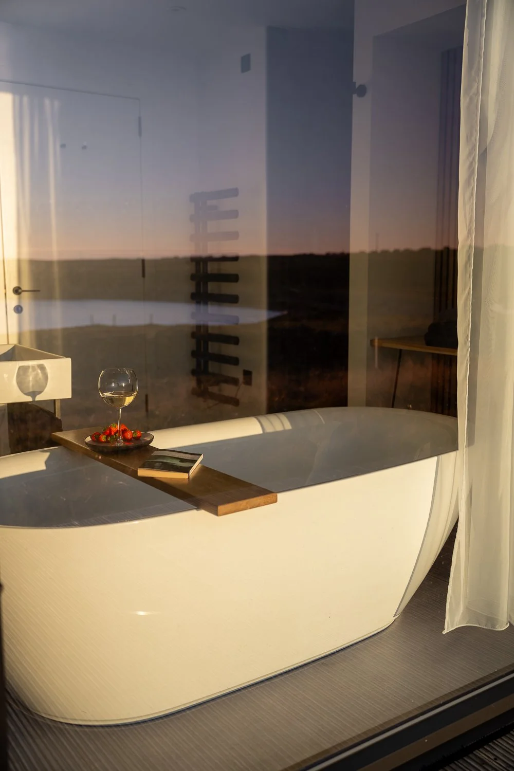 A tranquil view through a large window, revealing a relaxing bath positioned beside it. The bath features a caddy with a glass of wine, fresh strawberries, and an open book, creating an inviting atmosphere for a luxurious and peaceful retreat. 