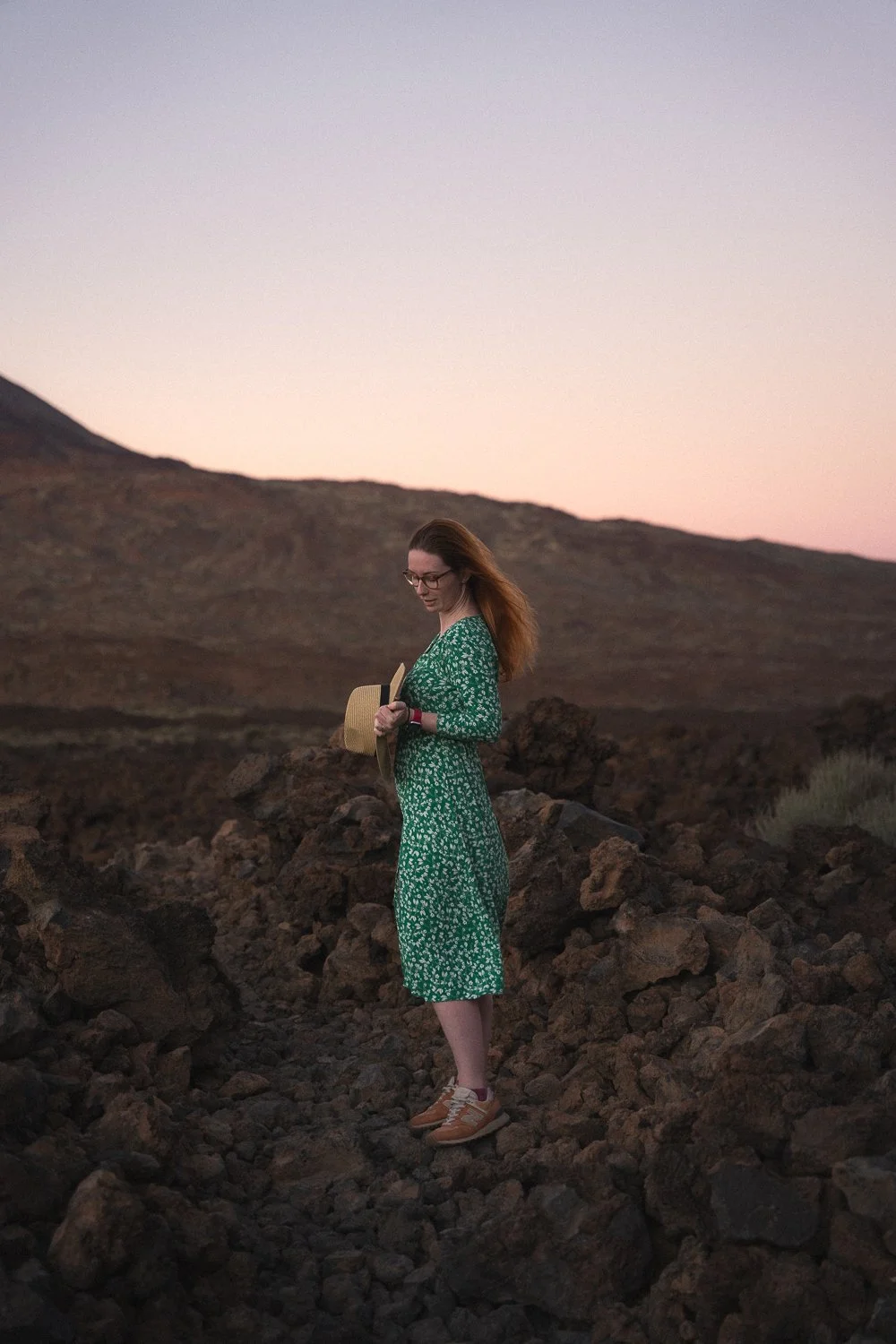 model in green dress standing in lava field holding hat.