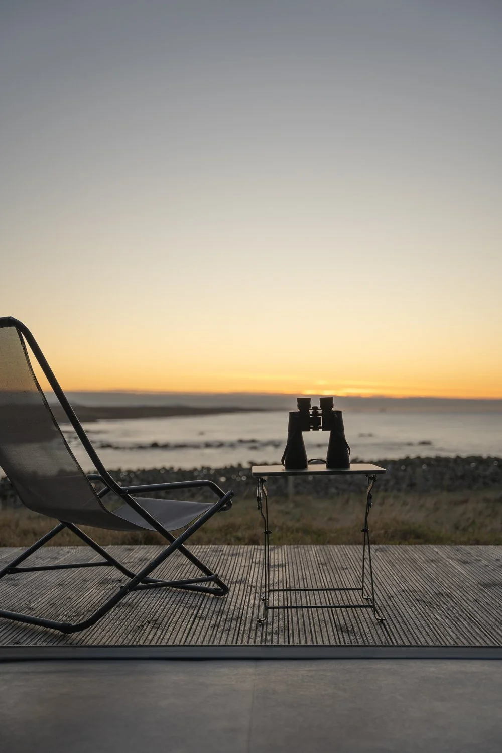 A captivating view from the house, featuring a deck with a chair and table adorned with binoculars. The scene captures the tranquil ocean just after sunset, with soft hues of yellow and orange reflecting on the water. 