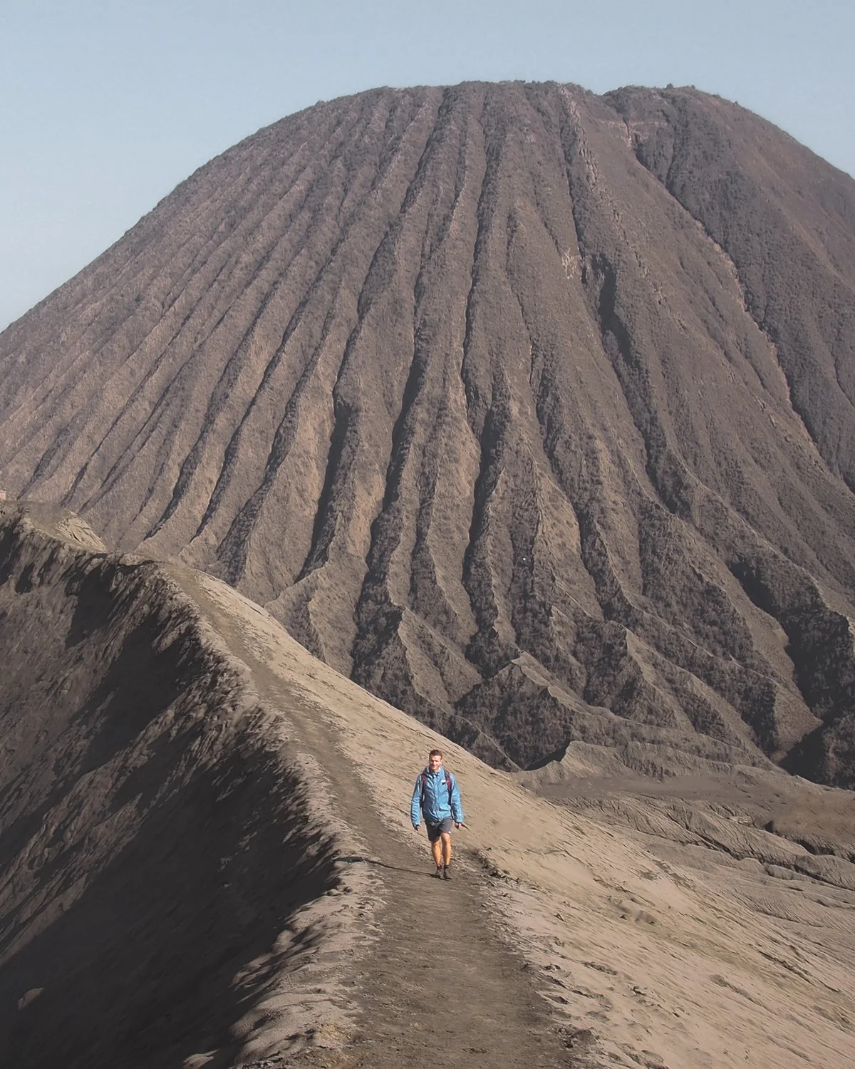 A person walking along volcano ridge with another volcano behind.
