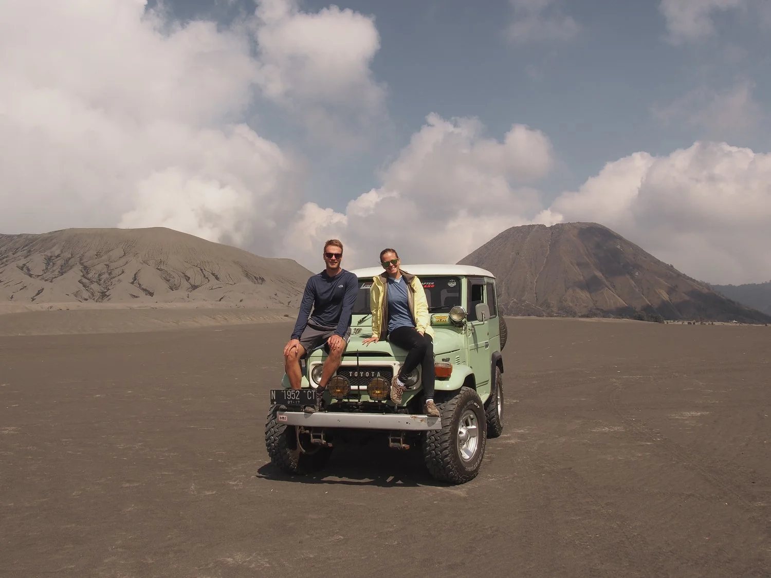 A couple sitting on the top of Jeep's hood which is parked in a crater with volcanoes behind.