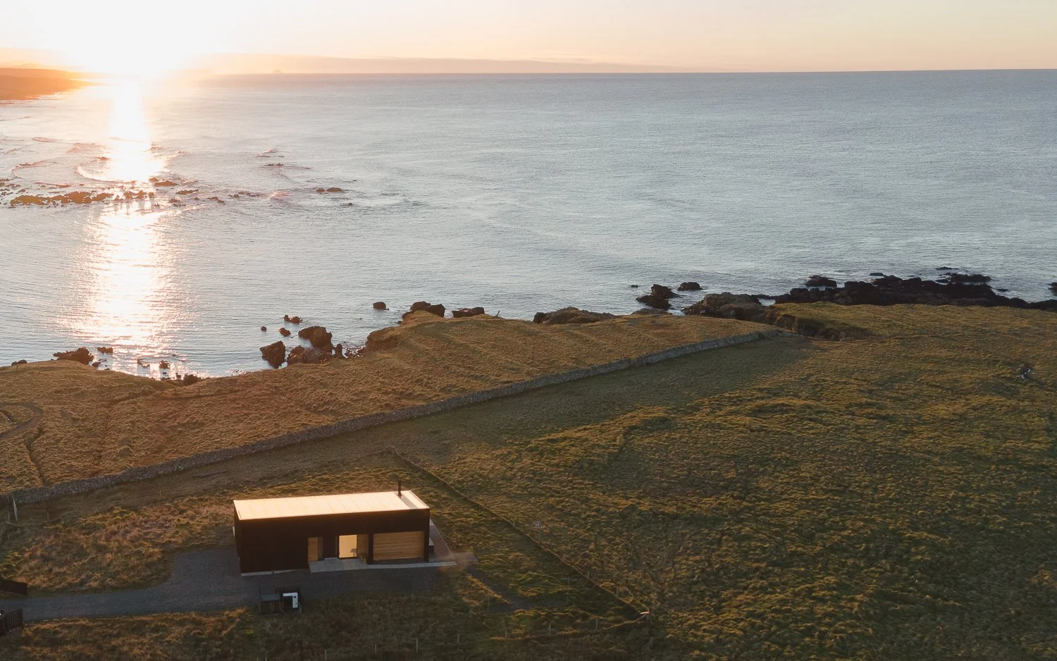 A breathtaking view of Brighid holiday home during sunset, bathed in golden light. The house sits elegantly on the edge of the ocean, surrounded by the stunning beauty of nature. Warm hues from the setting sun reflect off the water, creating a tranqu