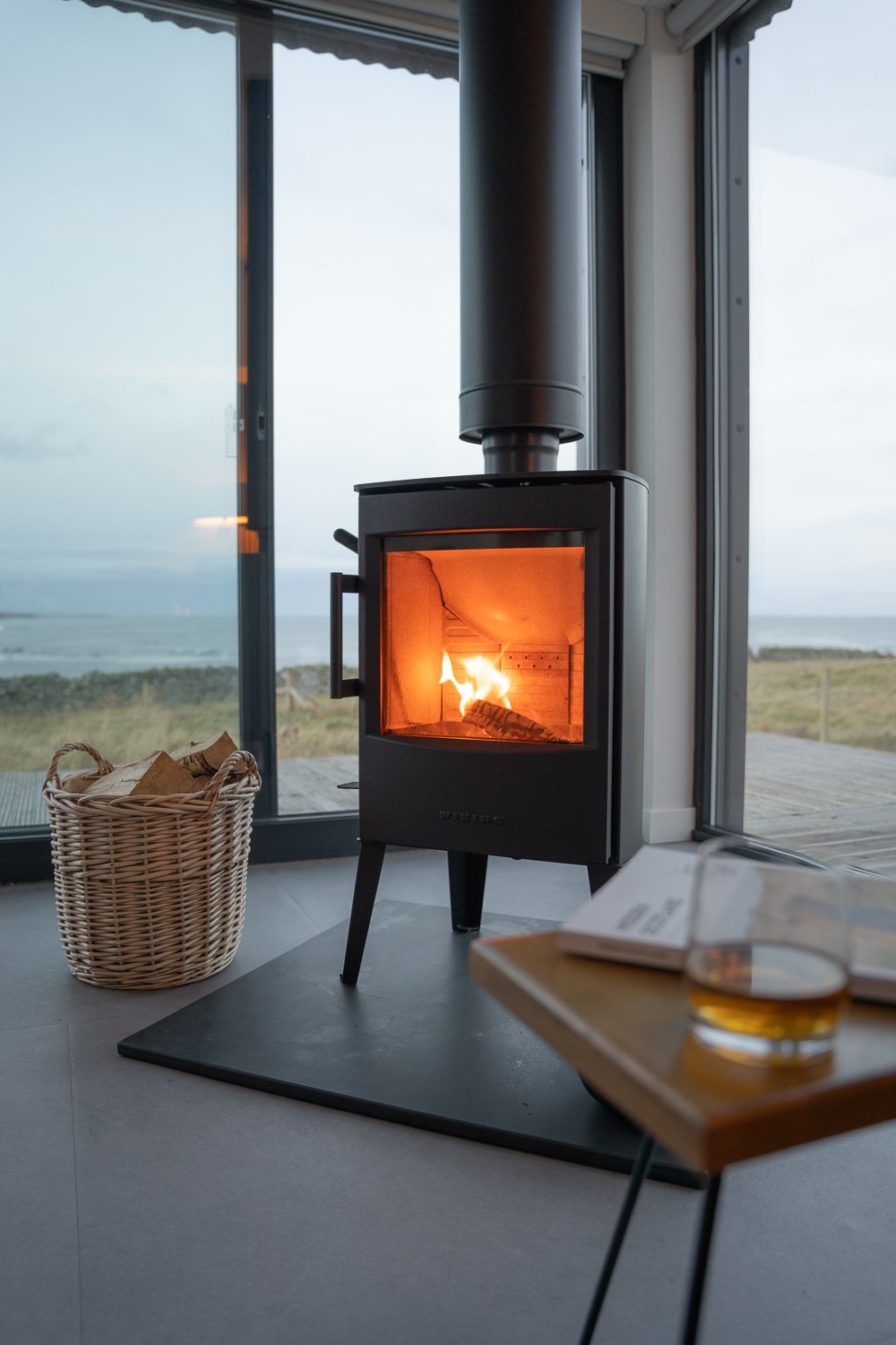 A close-up of a cozy living room fireplace, with a small coffee table in the foreground holding a book and a glass of whiskey. Beside the fireplace, a basket filled with firewood adds warmth and charm, while floor-to-ceiling windows in the background