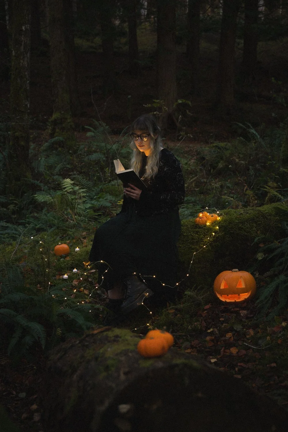 Model in black dress sitting down reading book in forest surrounded by fairy lights and carved pumpkins.