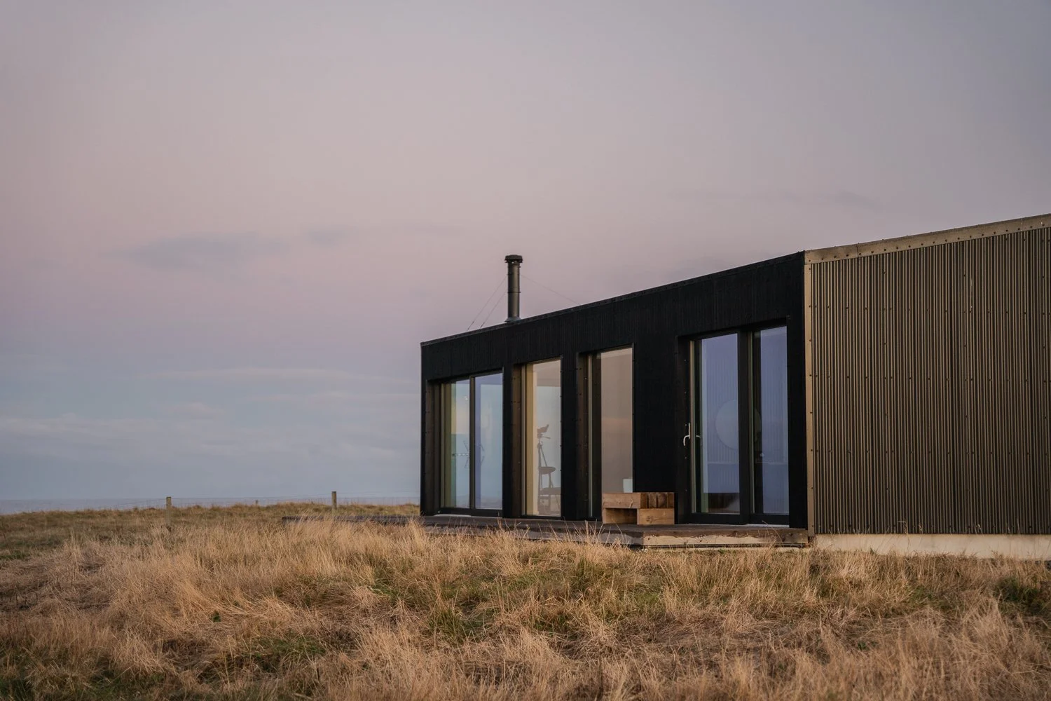 Modern black exterior of Brighid house on the Isle of Lewis, Scotland, during sunset. The building features large glass windows offering stunning views of the surrounding landscape, with dry grass and an open field in the foreground.