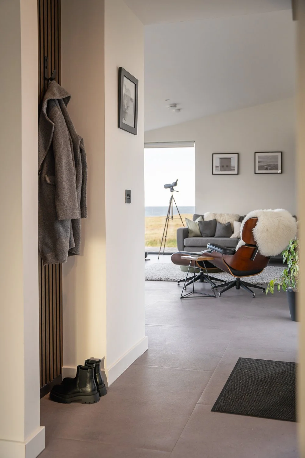 A cozy hallway featuring a hanging coat and shoes, leading into a welcoming living room in the background. The living area showcases a comfortable sofa, an inviting armchair, and a telescope, all framed by large windows that invite natural light. 