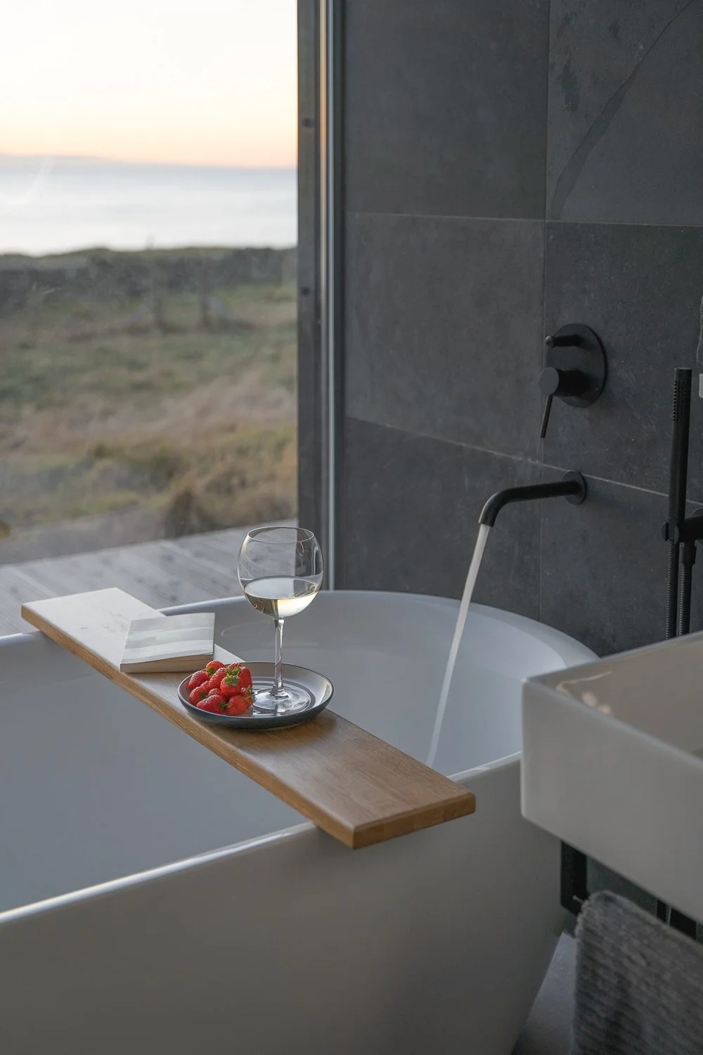 A serene bath with a running tap, featuring a caddy that holds a glass of wine, fresh strawberries, and an open book. This inviting scene creates a perfect atmosphere for relaxation and indulgence.