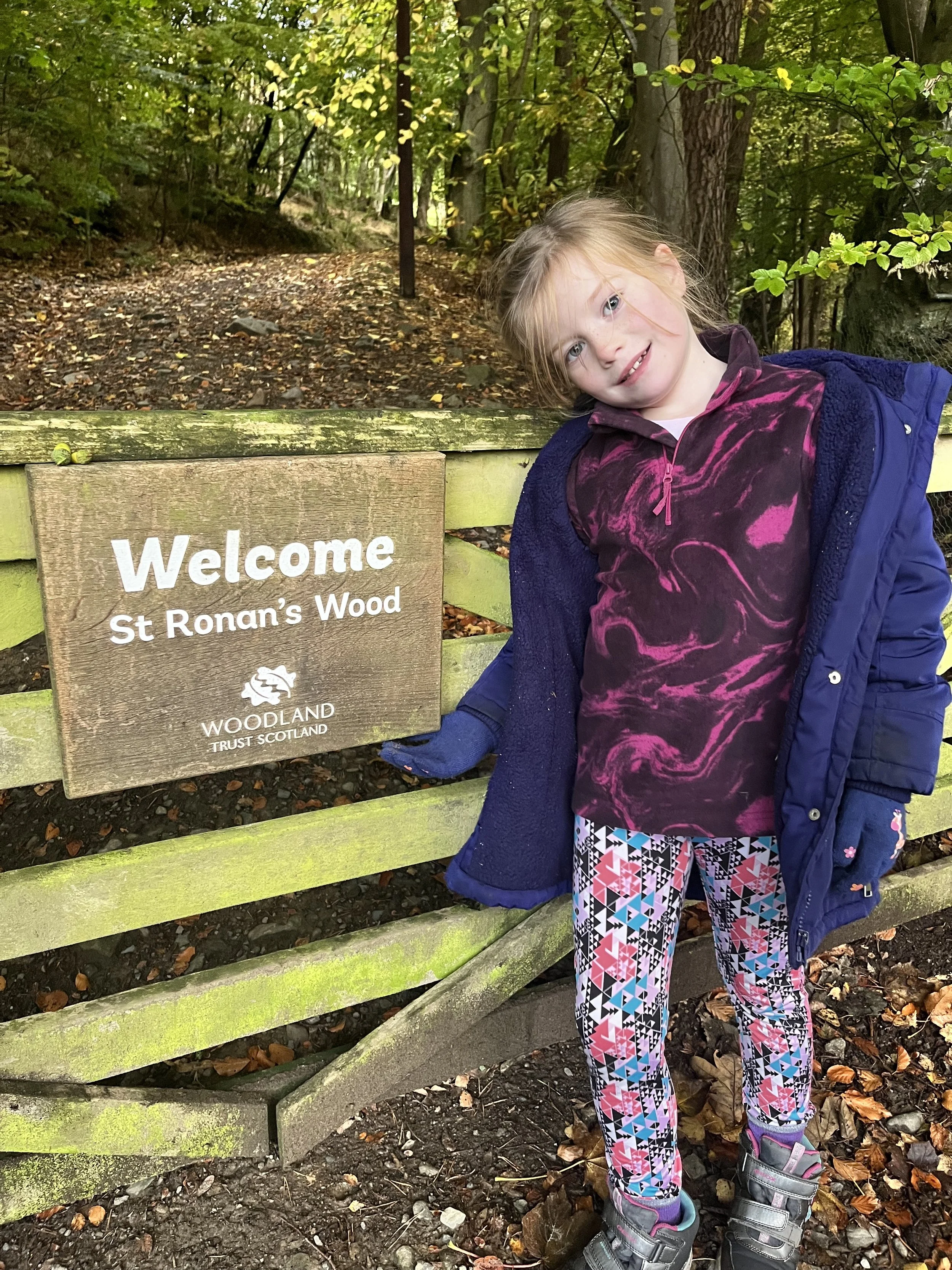 A young girl with light hair, wearing colorful patterned leggings, a purple marbled top, and a blue jacket, stands next to a wooden sign that says "Welcome St Ronan's Wood" in a forest setting.