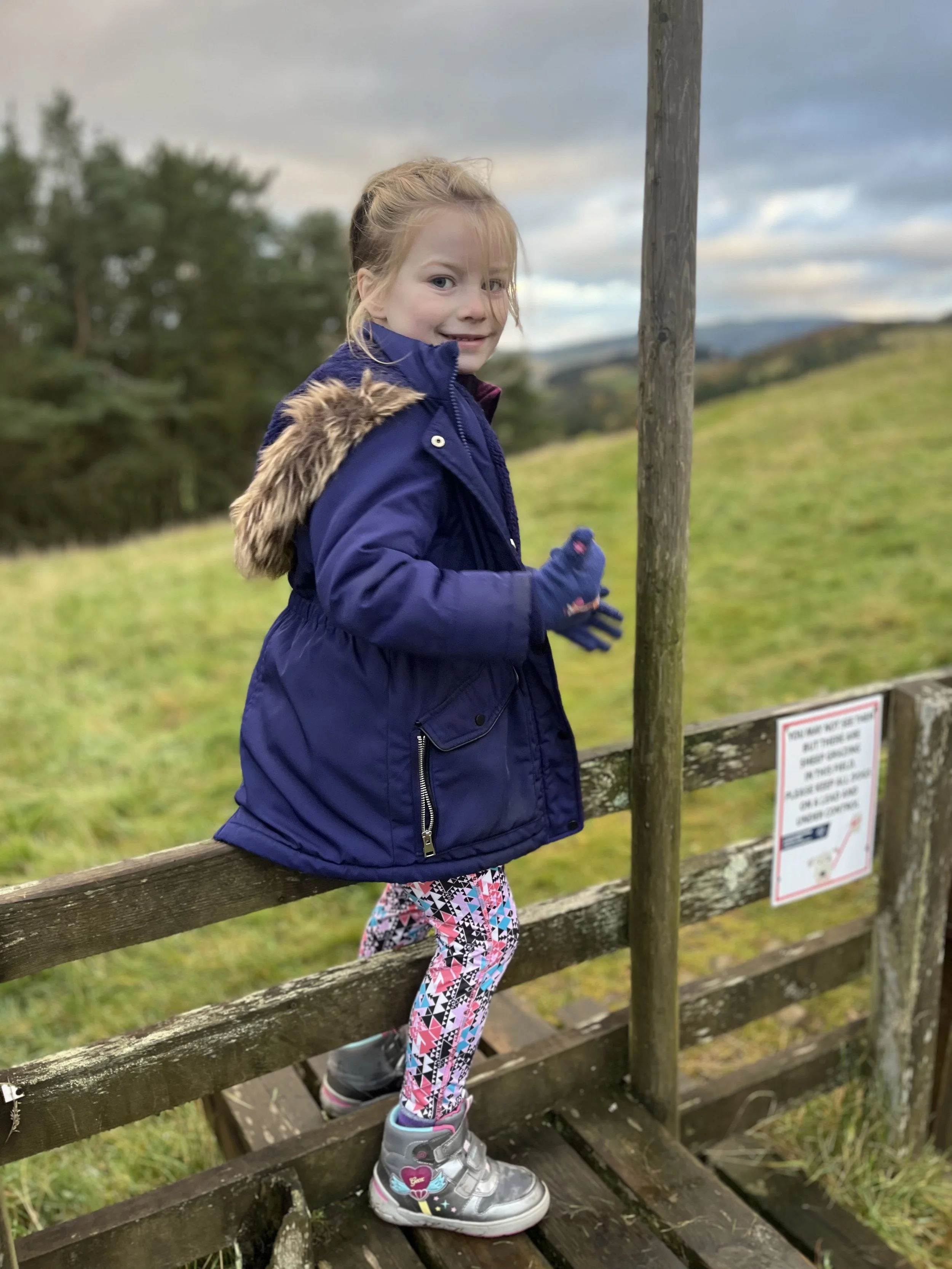 Young girl standing on a wooden bridge outdoors, wearing a dark blue coat with a fur-lined hood, colorful leggings, and gray boots, smiling at the camera with a scenic landscape in the background.