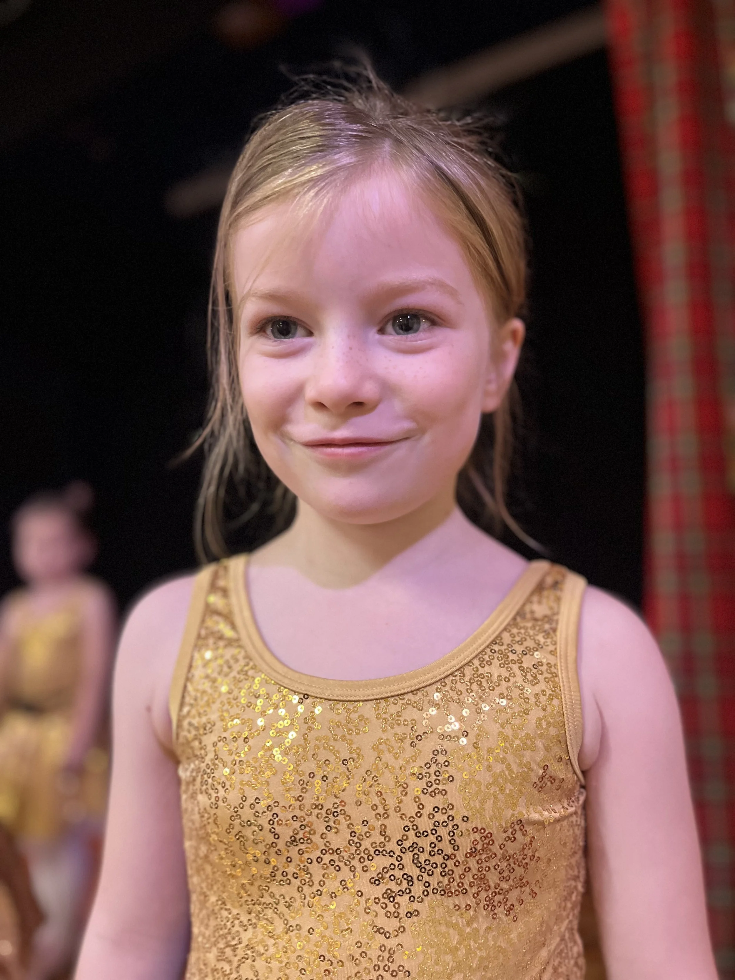 A young girl with red hair and freckles is smiling and wearing a gold sequined dance costume, standing in a dressing room.