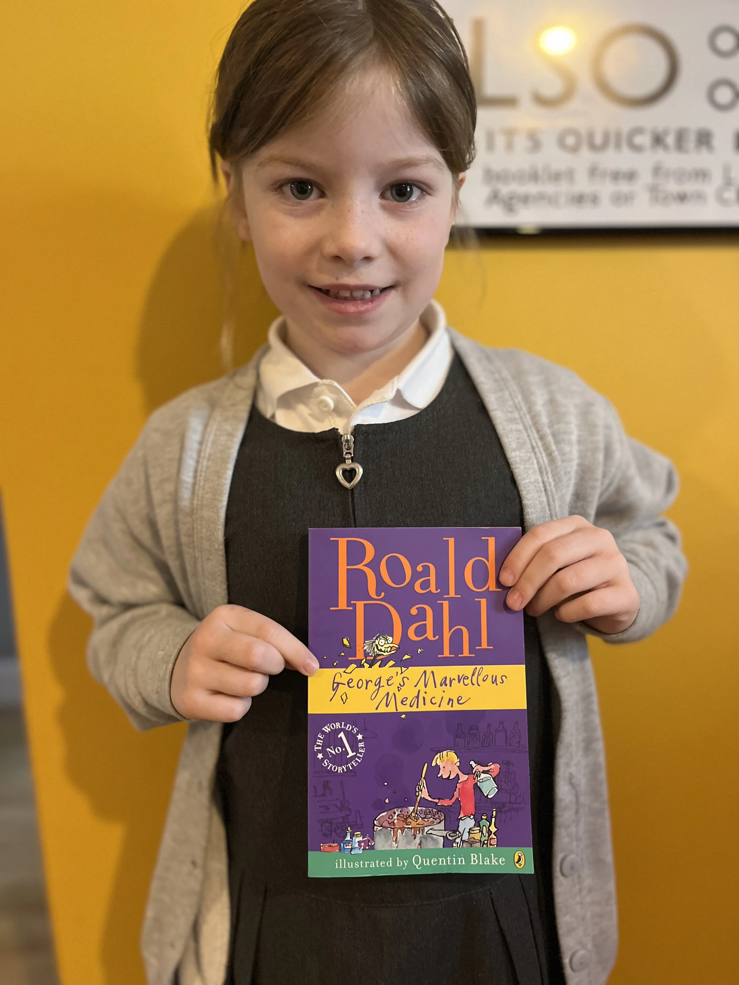 A young girl with brown hair, wearing a gray cardigan over a black dress and a white collared shirt, smiling while holding a purple book titled 'George's Marvellous Medicine' by Roald Dahl, with a yellow band and an illustration of a girl stirring a large pot on the cover. She is standing against a bright yellow wall.