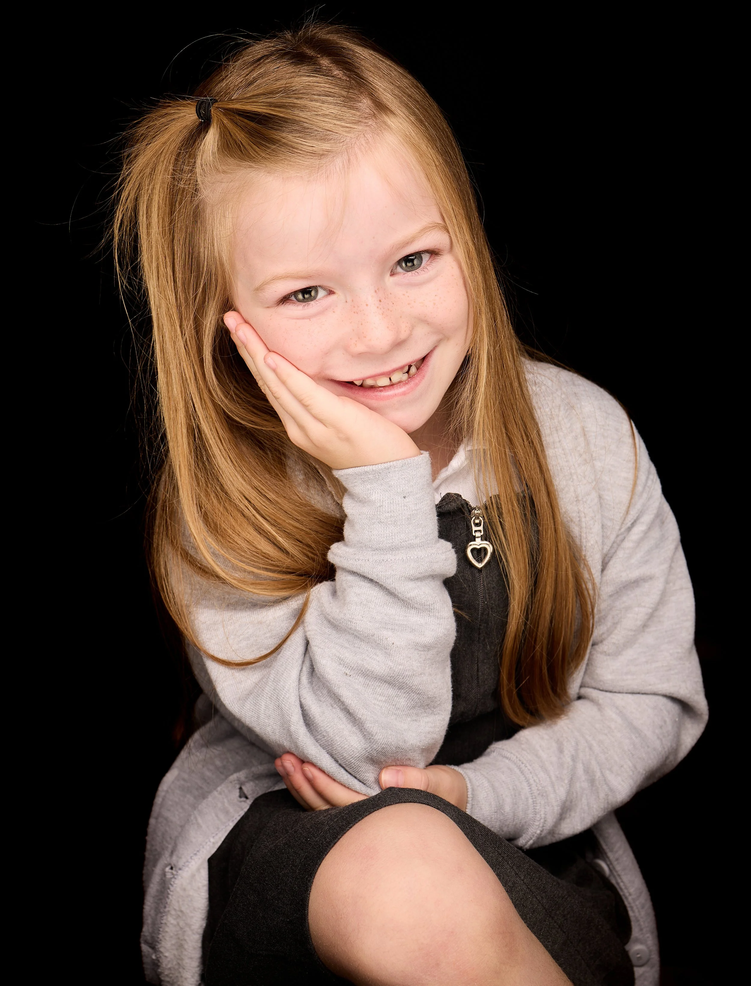 A young girl with red hair, freckles, and a big smile, sitting against a black background, resting her face on her right hand.