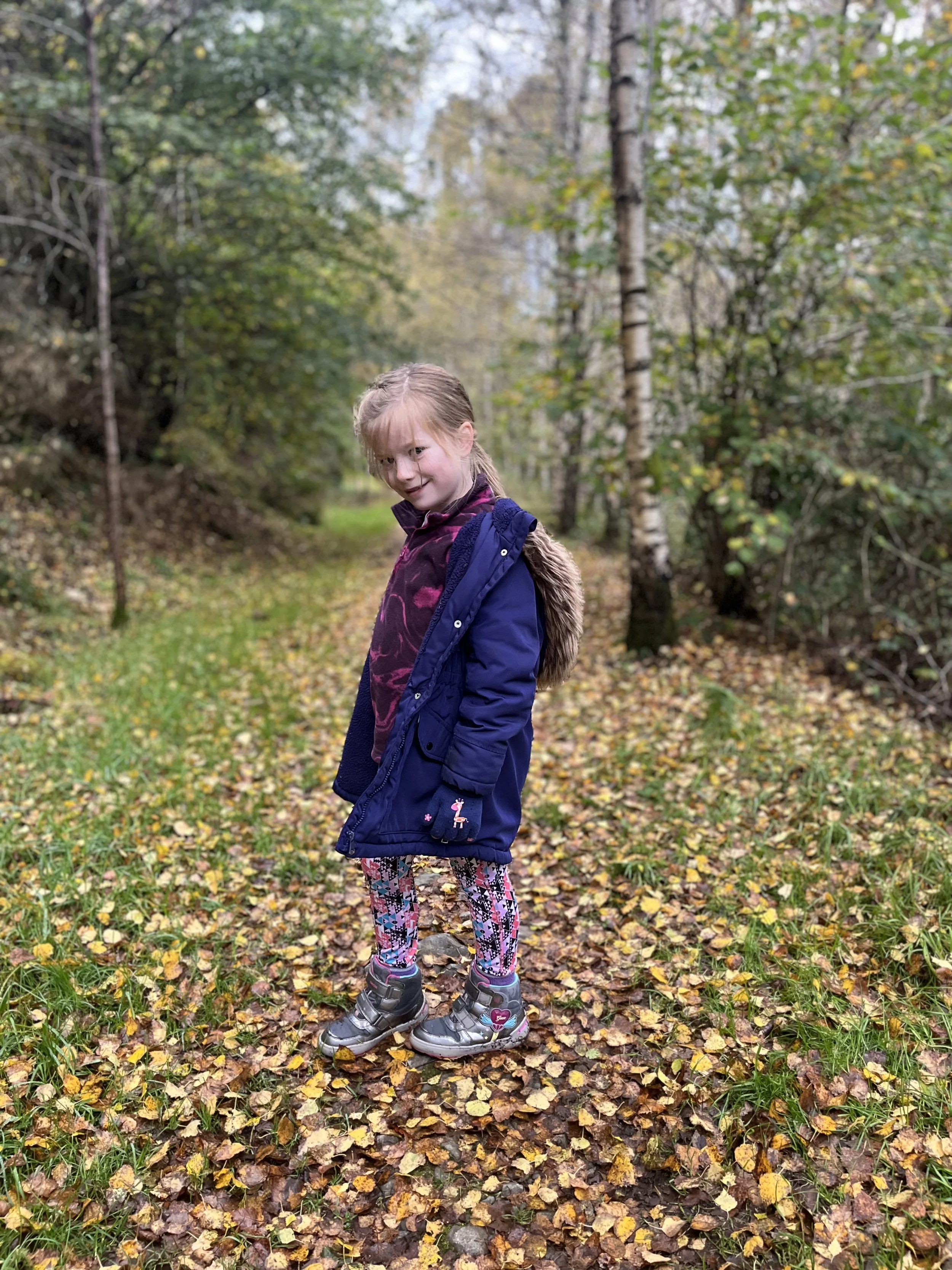 A young girl with blonde hair in a ponytail standing on a leaf-covered trail in a wooded area during fall, wearing a navy blue coat, patterned leggings, and hiking boots.