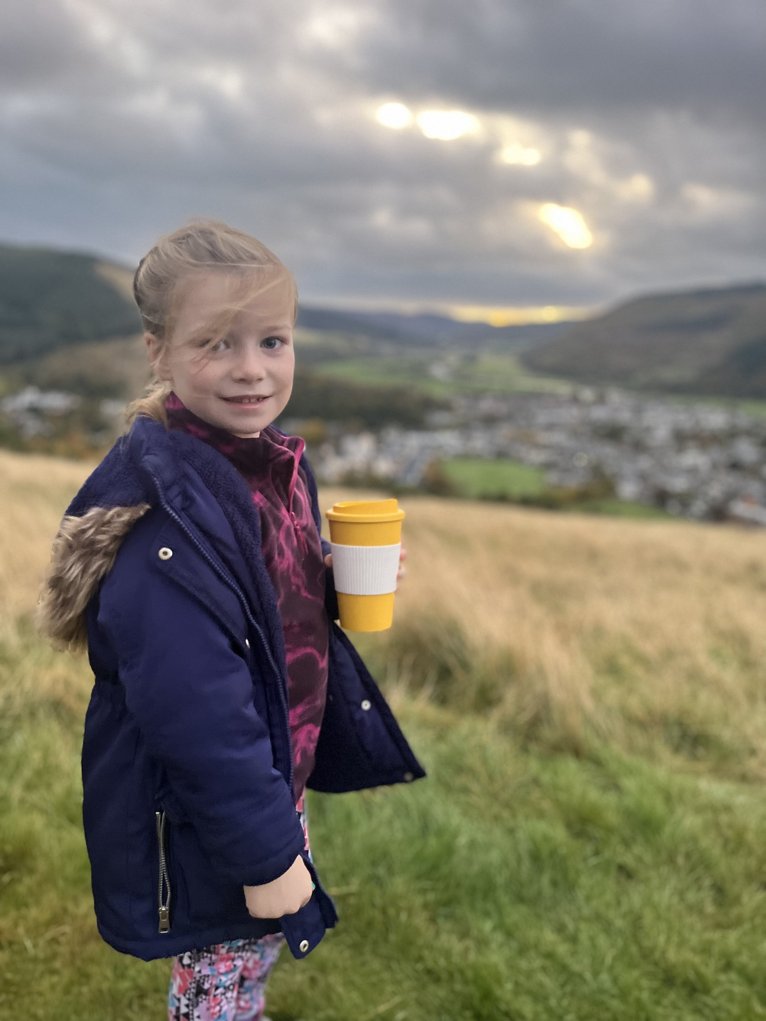 A young girl with blonde hair in a braid, wearing a dark blue jacket, floral pants, and holding a yellow and white travel mug, standing outdoors on a hill with a town and rolling hills in the background under a cloudy sky at sunset.