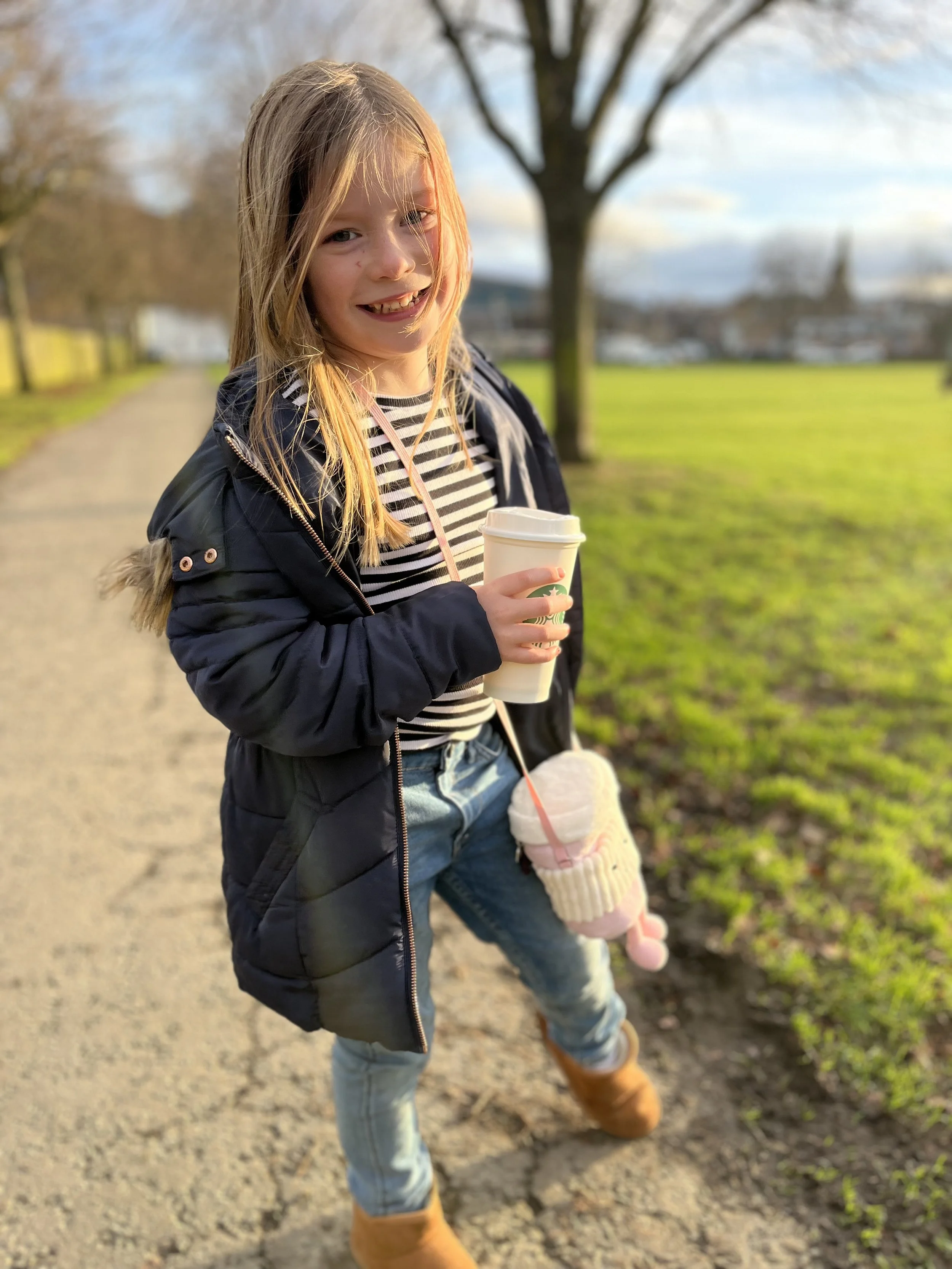 A young girl with long blonde hair standing outdoors on a path, holding a cup of a hot drink, dressed in a black jacket, striped shirt, jeans, and tan boots, with a pink plush toy hanging from her pocket, and smiling at the camera.