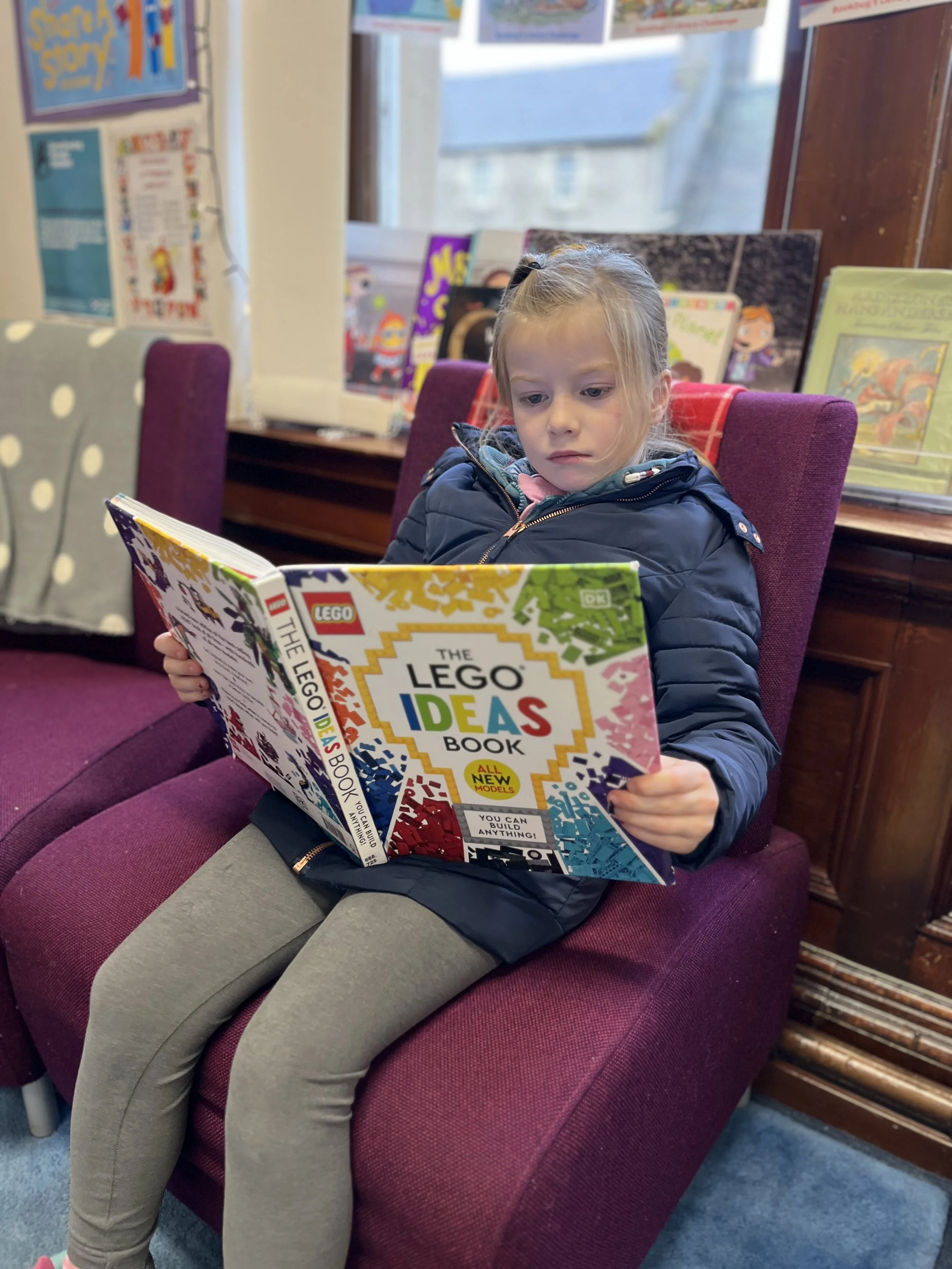 Young girl sitting in a deep purple armchair reading a colorful book titled "The LEGO Ideas Book" inside a bookstore or library, with shelves of books and a window in the background.