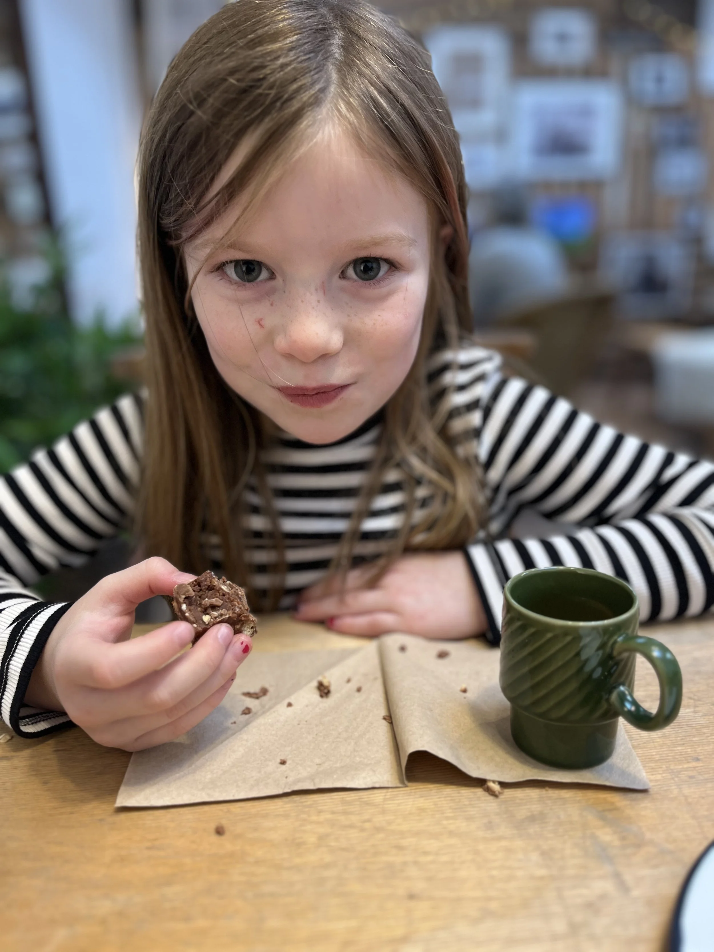 A young girl with long brown hair and freckles sitting at a wooden table, holding a piece of chocolate cookie in her hand, next to a green mug with a hot beverage, in a cozy indoor setting with photos on the wall in the background.