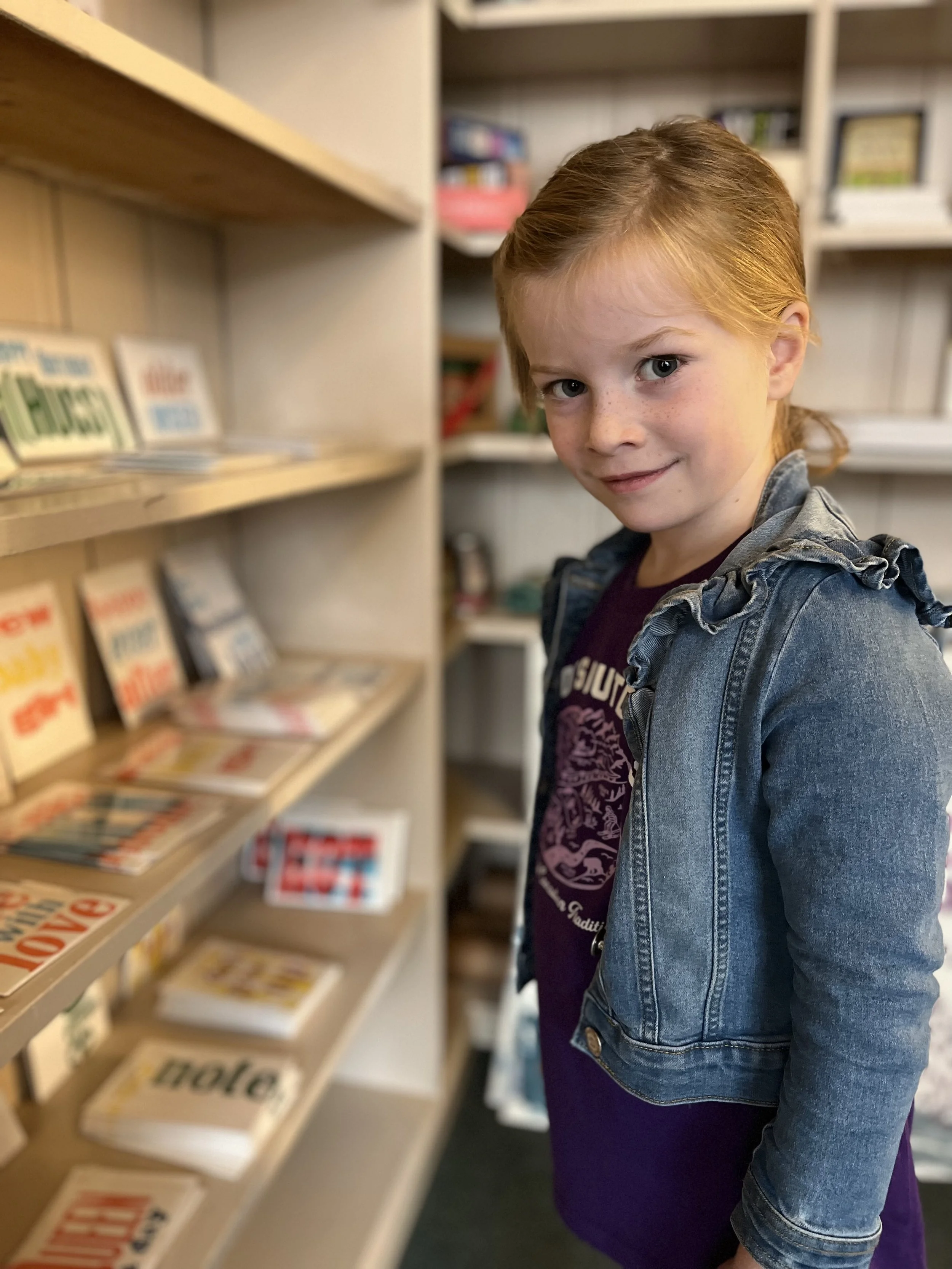 A young girl with red hair and freckles standing in front of a bookshelf filled with books, wearing a denim jacket and a purple shirt with a graphic design.