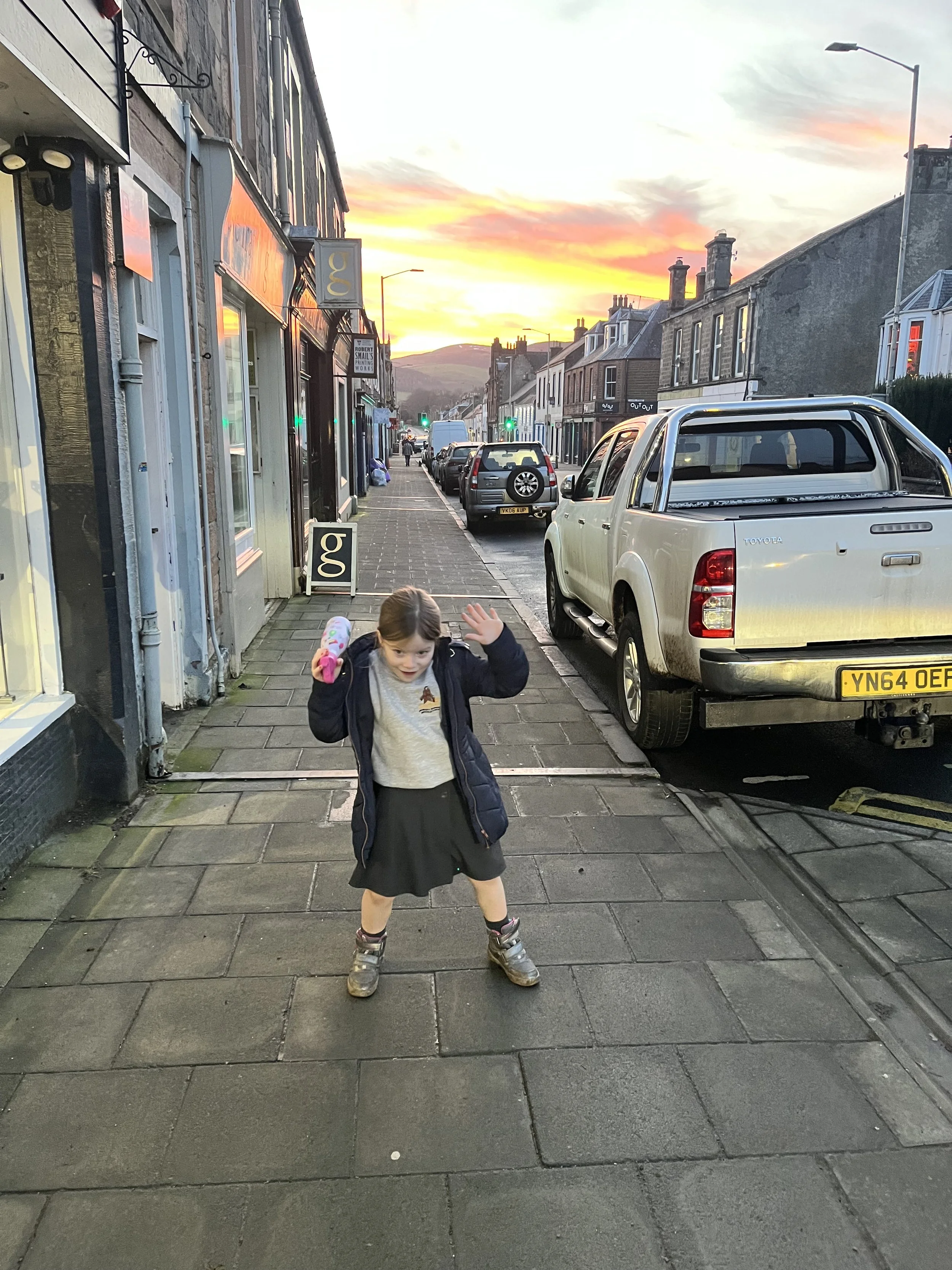 A young girl wearing a school uniform, jacket, and sneakers, standing on a sidewalk at sunset, waving with a playful expression.