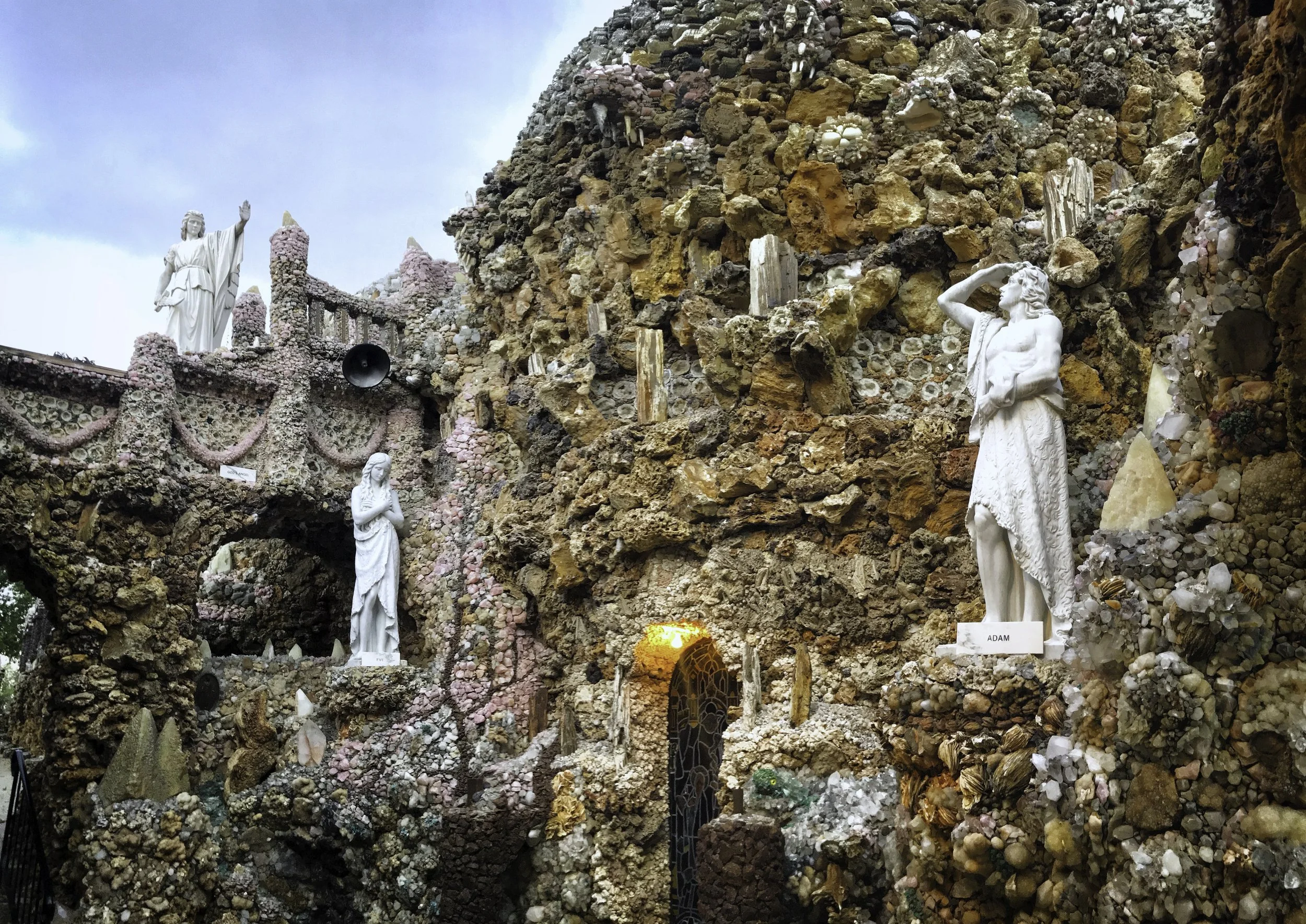 Sculptures of religious figures set against a textured, rocky background with colorful shells and stones, including statues of Jesus and Adam.