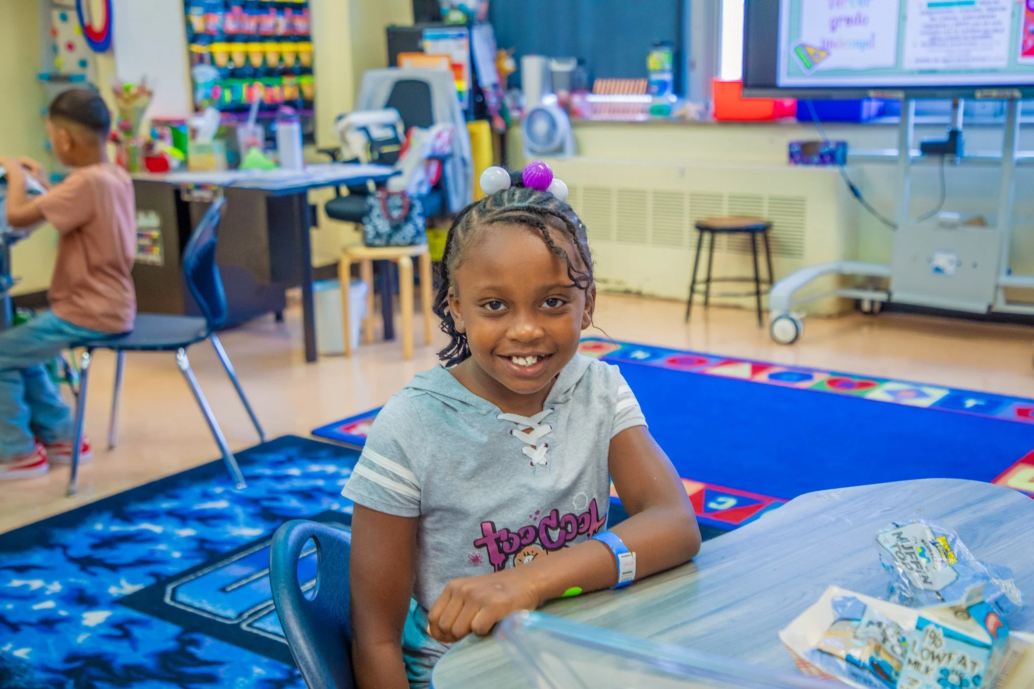 A young girl with braided hair and colorful beads in her hair, smiling at the camera, sitting at a desk in a classroom. In the background, a boy is sitting at another desk, with educational supplies and a large screen on the wall.