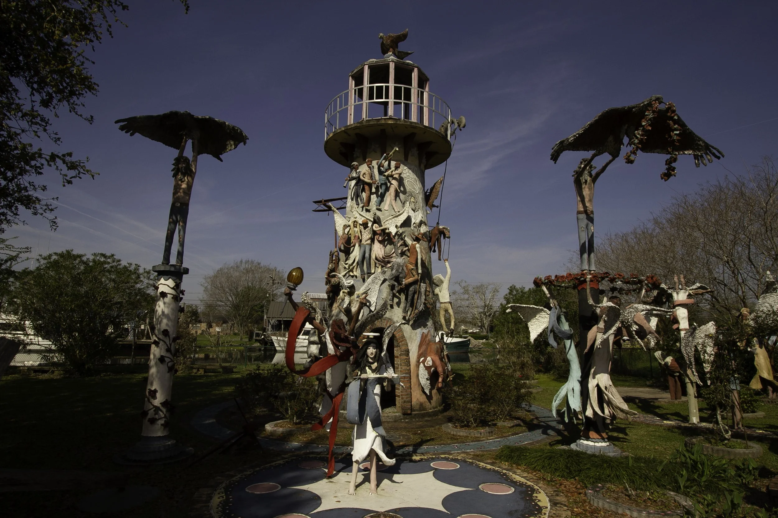 Colorful, eclectic outdoor sculpture featuring numerous human and animal figures, set against a clear blue sky, with trees and greenery around.