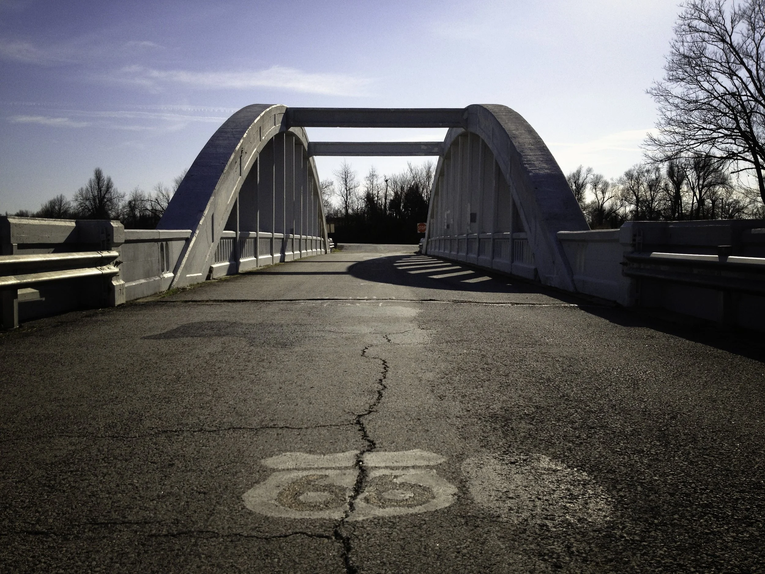 A small concrete bridge with white railings over a cracked asphalt road with a painted Route 66 symbol. Bare trees on the right side and a stop sign in the distance.