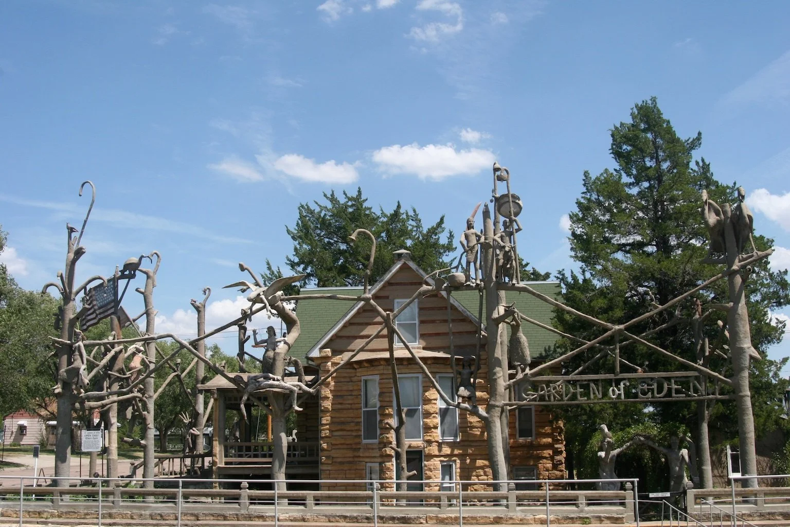 Wooden house with a green roof behind a metal fence, surrounded by leafless tree sculptures with human and animal figures, and a sign reading "Garden of G Dorm".