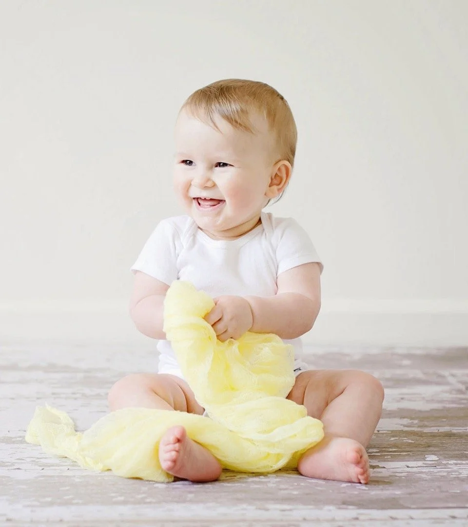A happy toddler with red hair sitting on a wooden floor, playing with yellow fabric, wearing a white shirt.