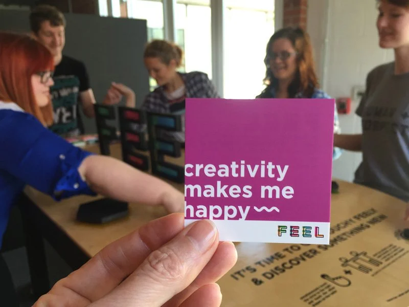 Student work: A person holds a purple card that reads 'creativity makes me happy' in front of a group of people indoors, with some of them smiling and engaging in conversation.