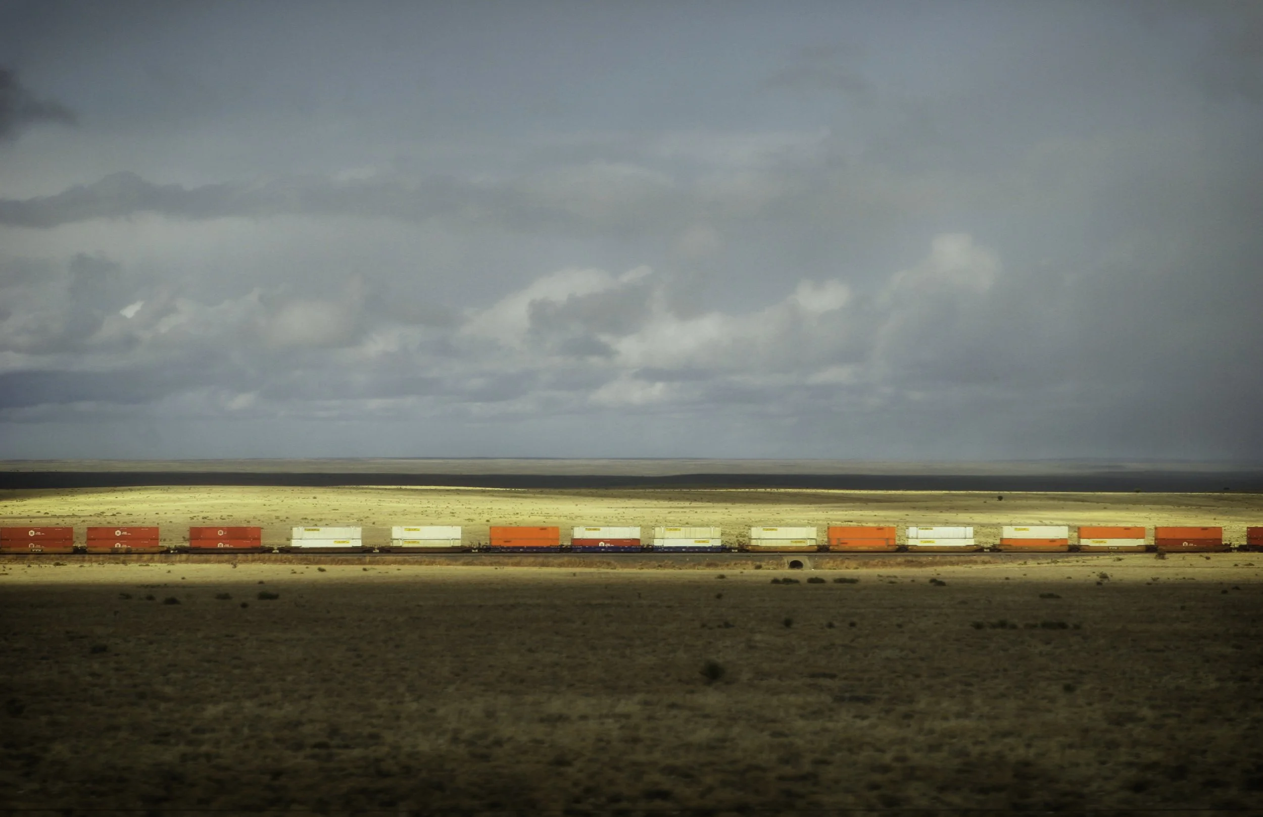 A train with red, white, and orange cargo containers traveling through a vast, flat desert landscape under a cloudy sky.