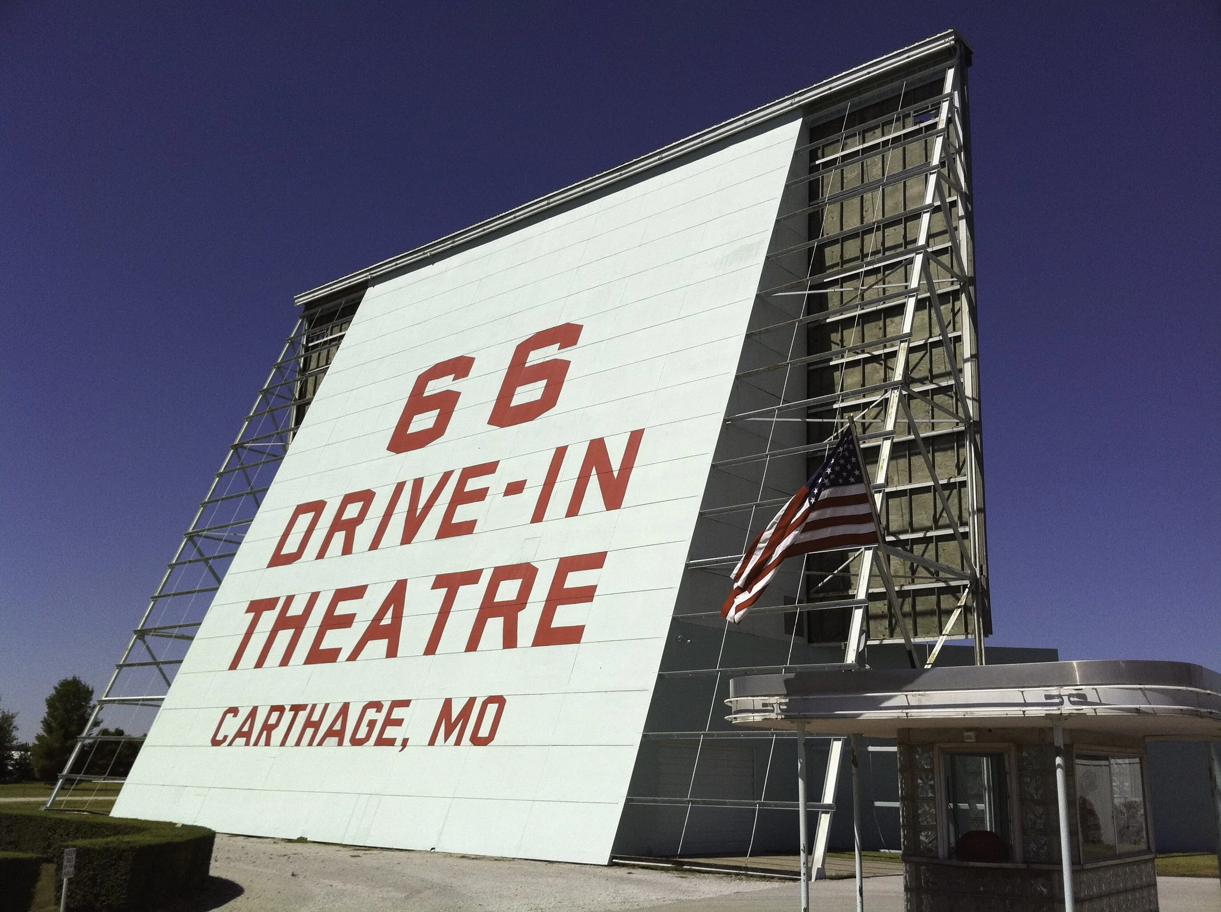 A vintage drive-in theater sign with a large white signboard with red text advertising a 66 drive-in theater in Carthage, Missouri. The sign has a metal frame, an American flag, and a small ticket booth in front.
