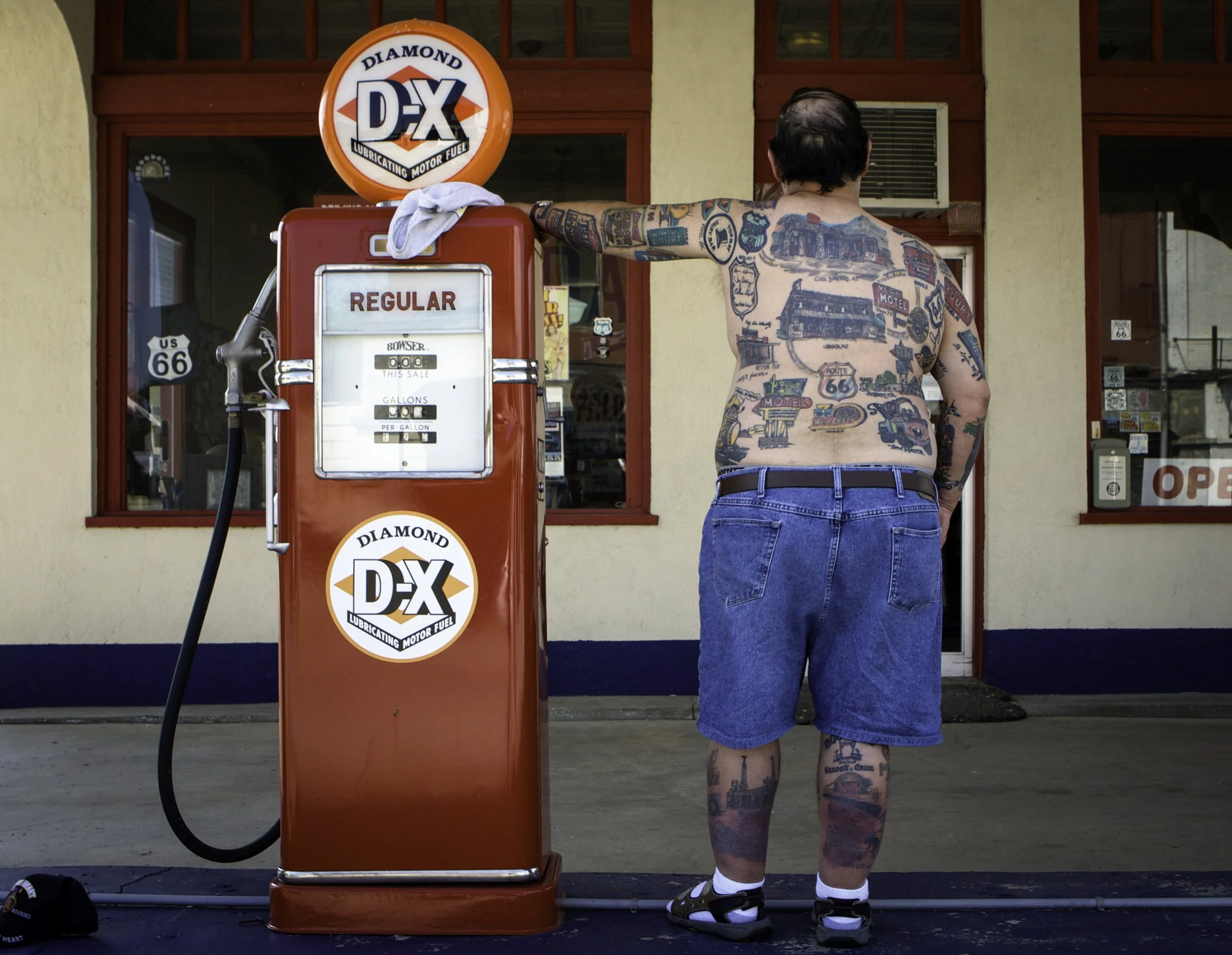 A shirtless man with tattoos standing at a vintage gas pump outside a store on Route 66.