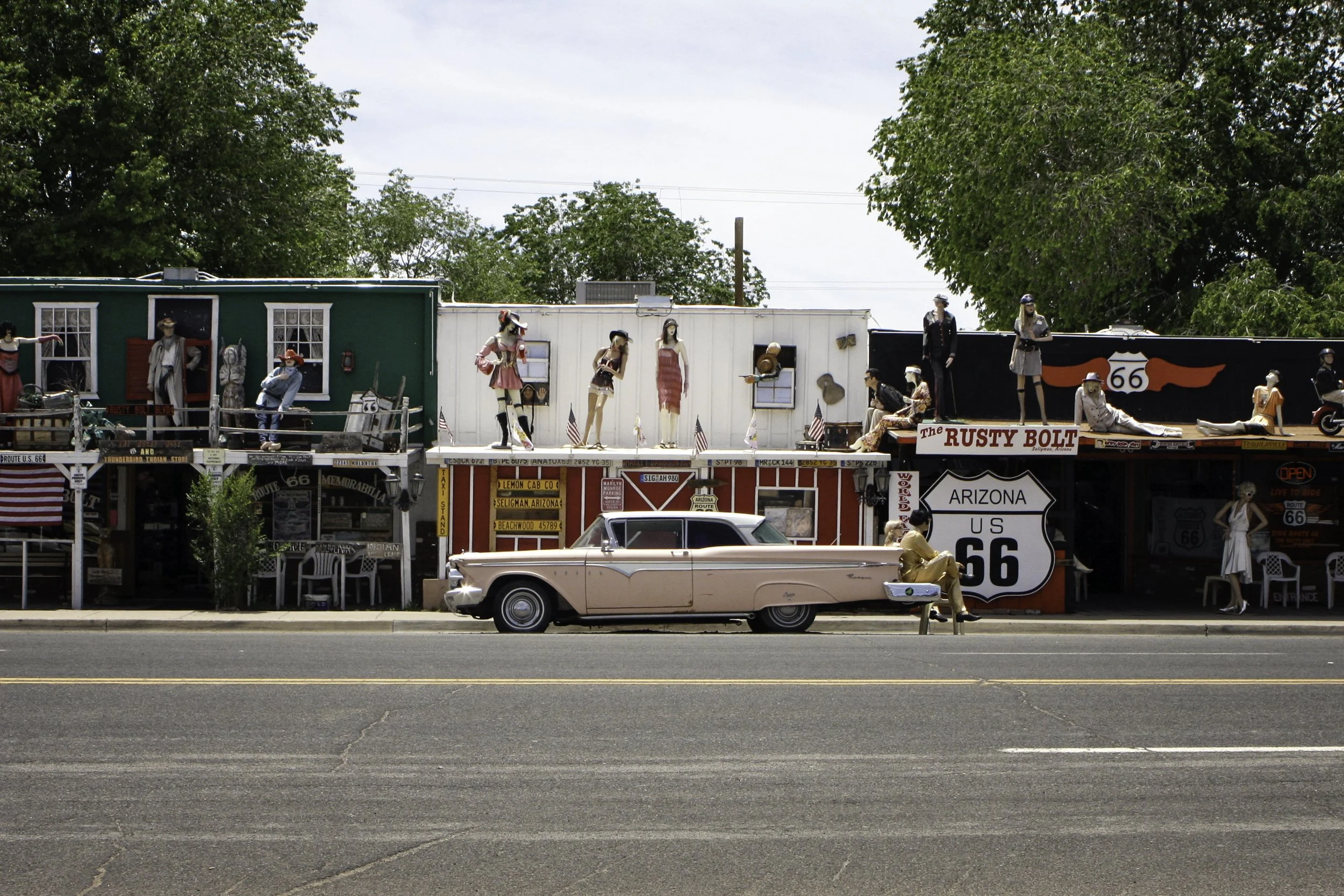 Colorful roadside scene with mannequins dressed in vintage clothing on a second-story balcony, a pink classic car parked in front, and Route 66 signs, all against a backdrop of green trees and clear blue sky.
