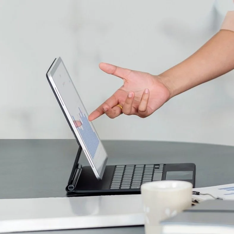 Person using a touchscreen tablet mounted on a portable stand over a laptop, with a coffee mug nearby.