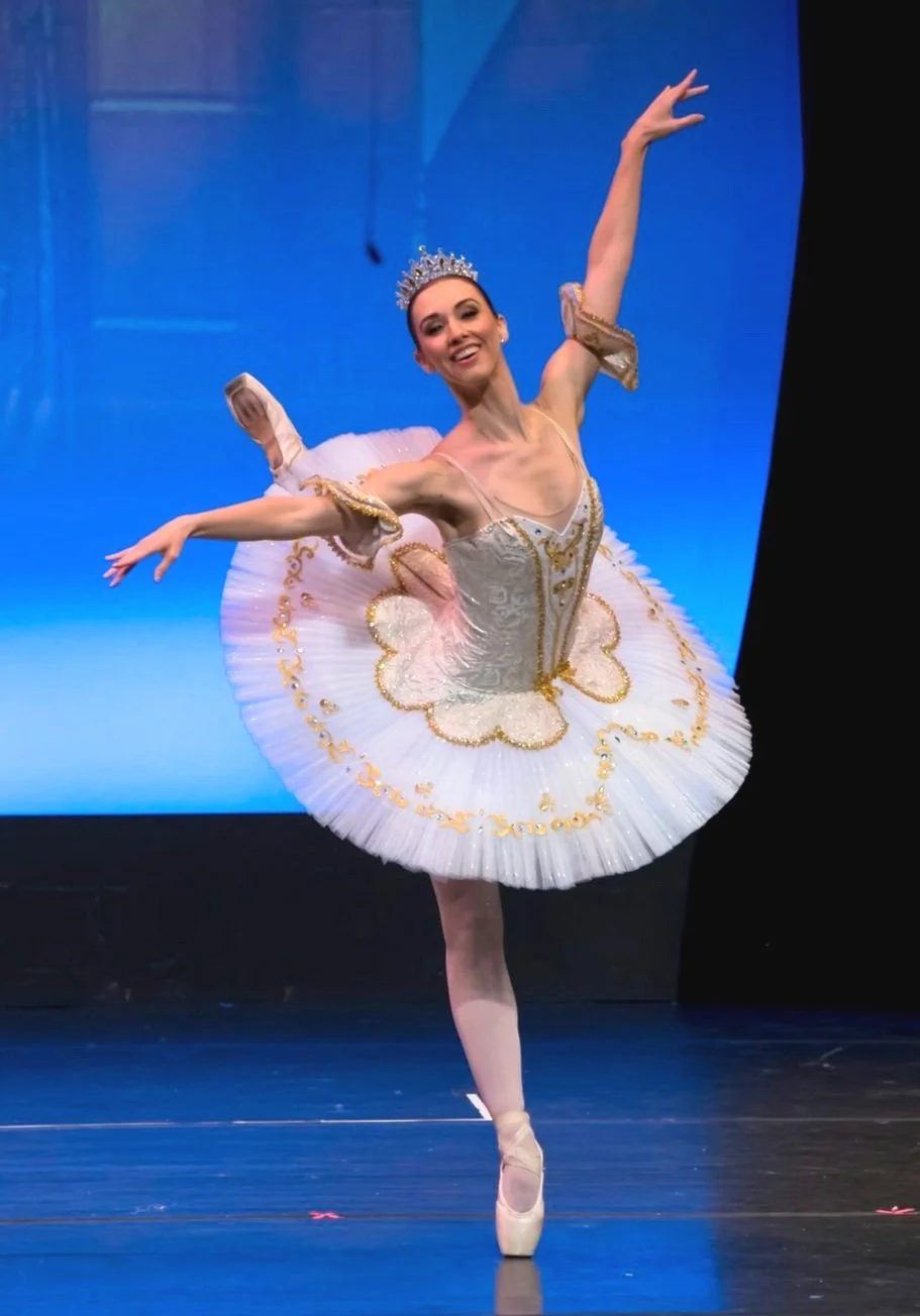 A ballet dancer with a crown performs a pose on stage, wearing a white and gold tutu and pointe shoes, with a blue background.