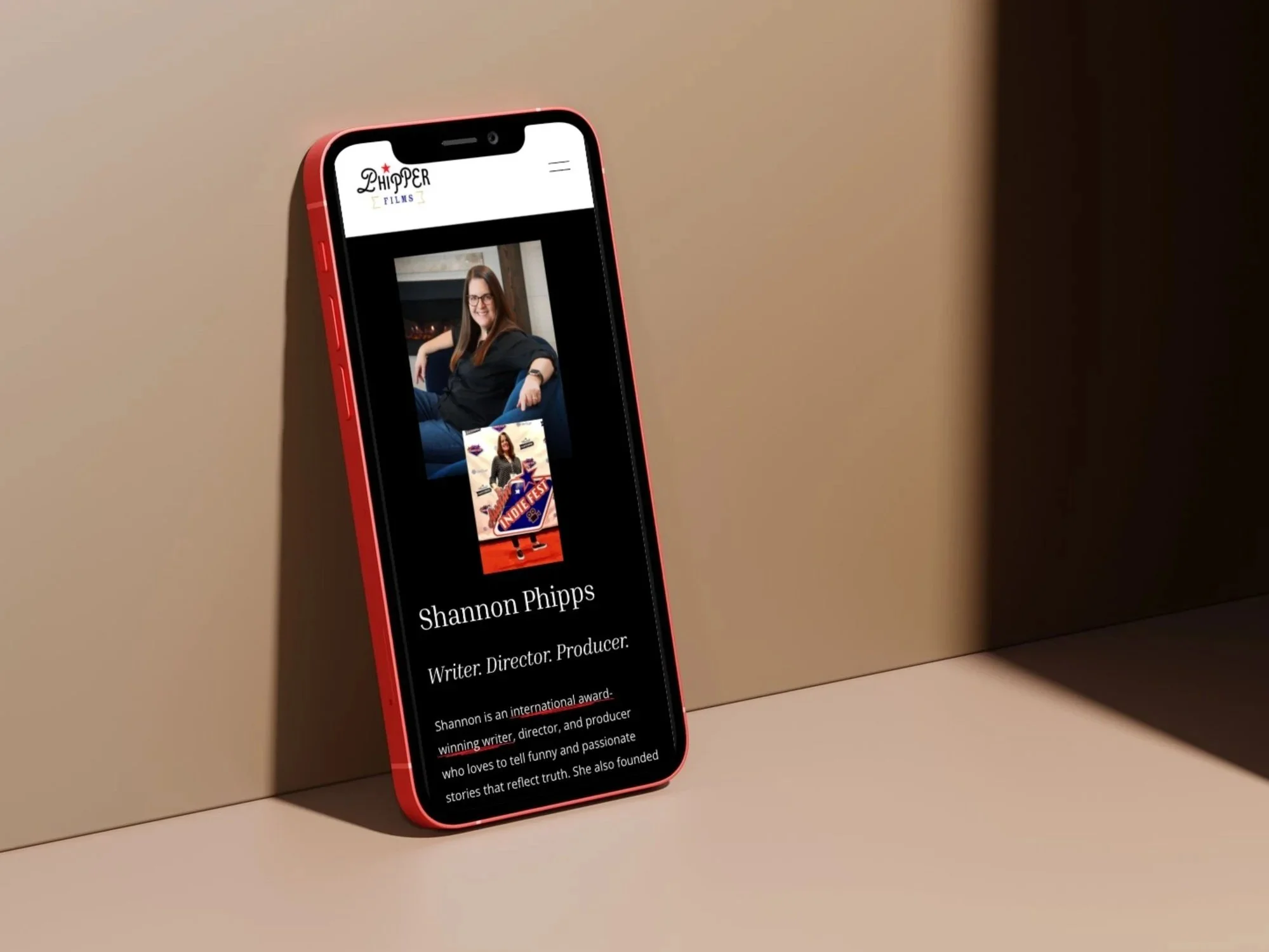 A red smartphone displaying a personal webpage of Shannon Phipps, a writer, director, producer, with a photo of her sitting on a blue couch and a poster of her work.