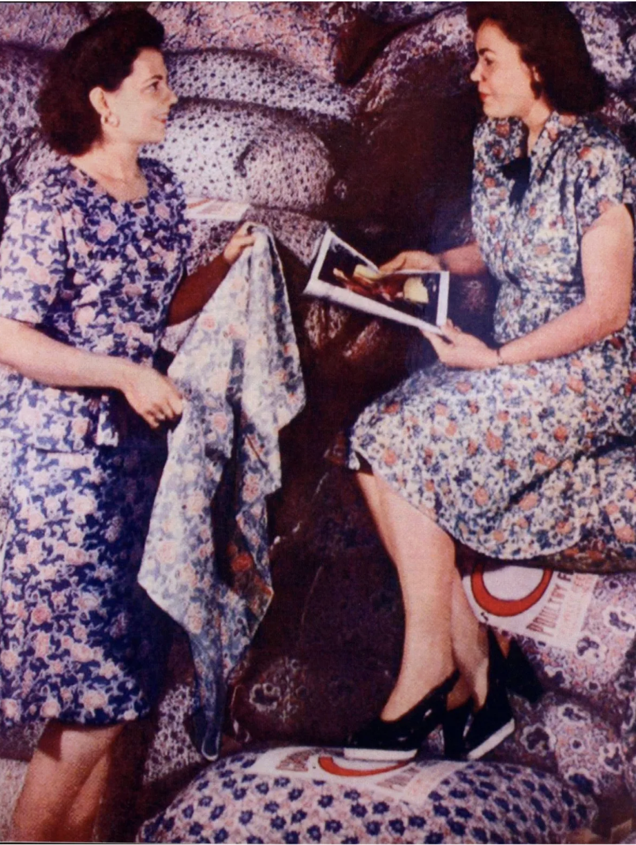 Two women in floral dresses sitting on patterned furniture, engaging in conversation, one holding a magazine, in a vintage setting.