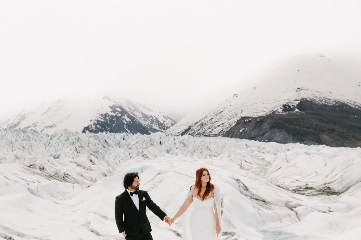 A bride and groom holding hands on a glacier with snow-covered mountains in the background.