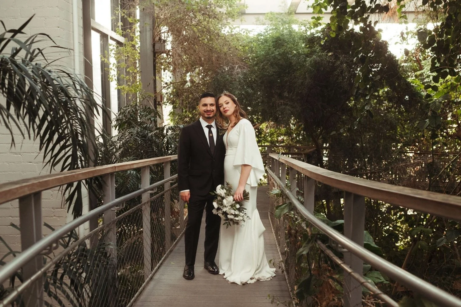 A bride and groom stand together on a wooden bridge inside the Texas Discovery Gardens greenhouse.