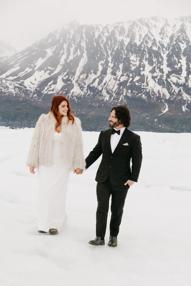 A couple dressed in wedding attire walking hand-in-hand in a snowy landscape with a rocky, snow-covered mountain in the background.