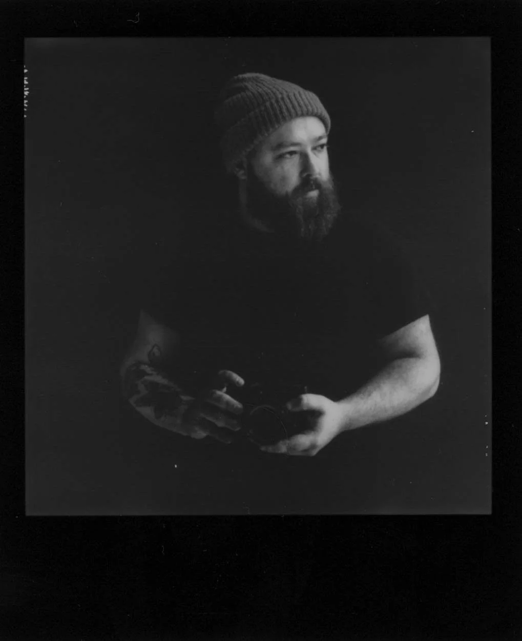 A bearded Dallas wedding photographer in a beanie holding a camera against a dark background.