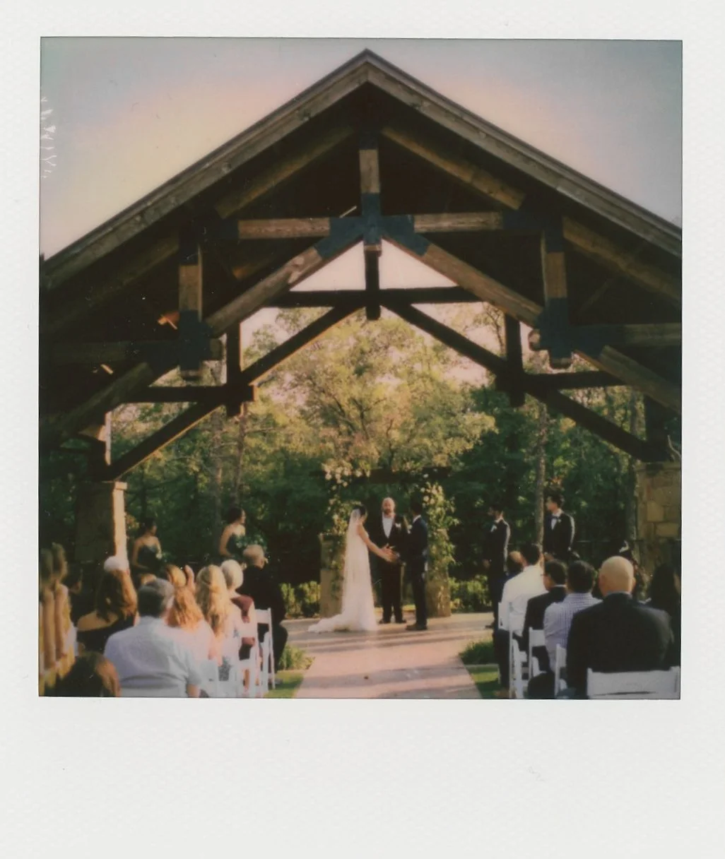 An Instax Square photo shows a bride and groom exchanging vows at an outdoor altar under a wooden pavilion as guests watch from their seats.