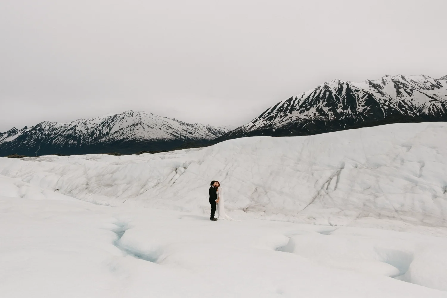A couple stands close together on a snowy, icy landscape with snow-covered mountains in the background.