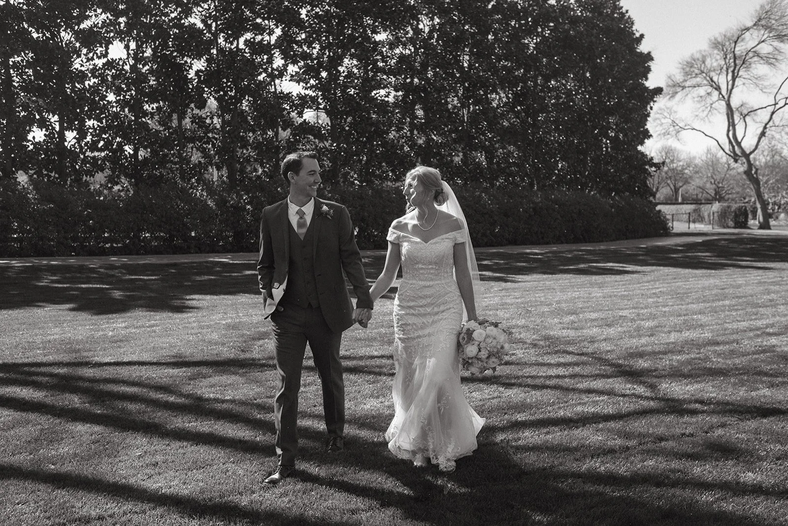 A bride and groom smile while walking hand in hand across the Dallas Arboretum lawn.