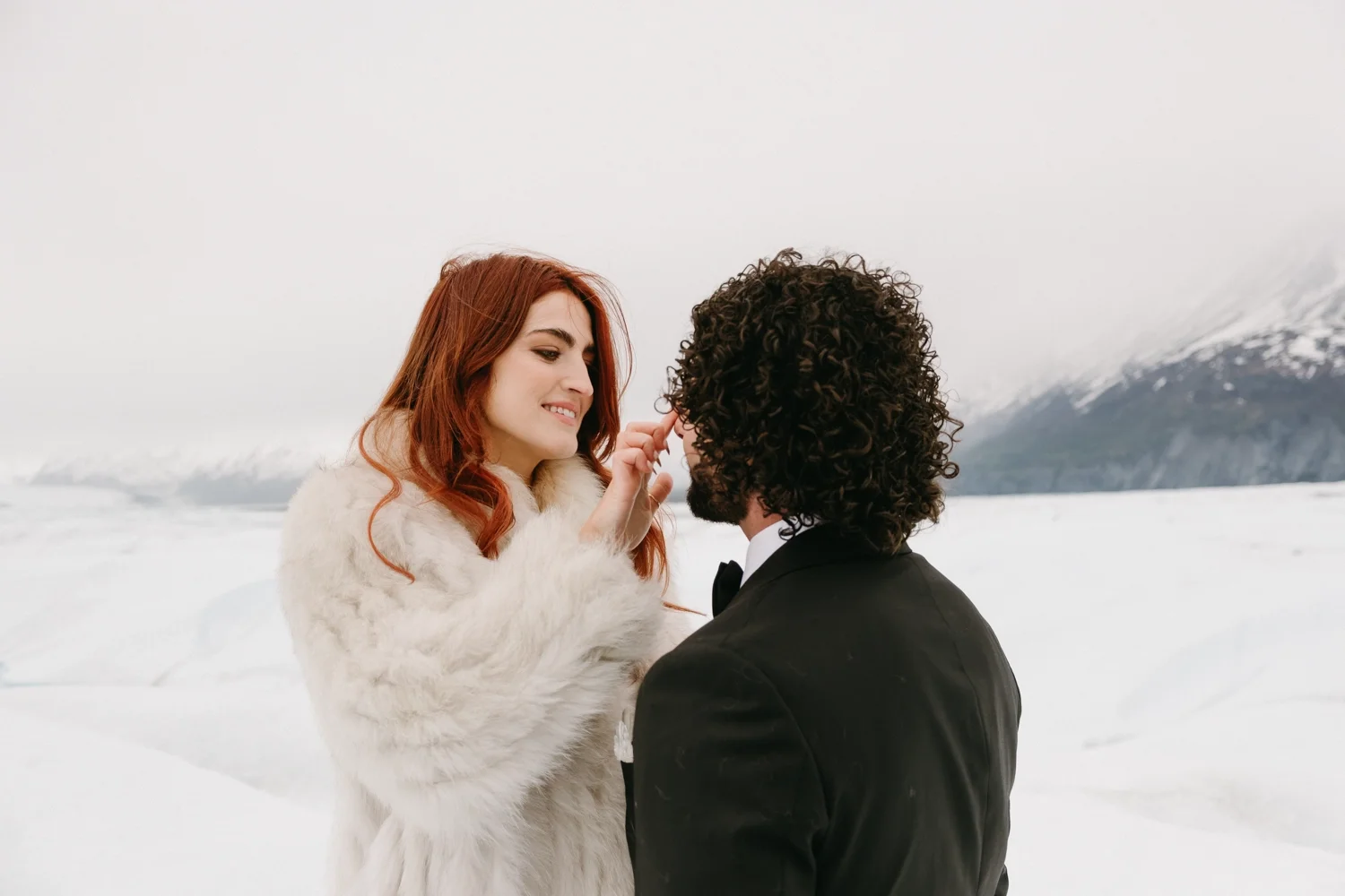 A couple dressed in formal attire sharing an intimate moment in a snowy mountain landscape.