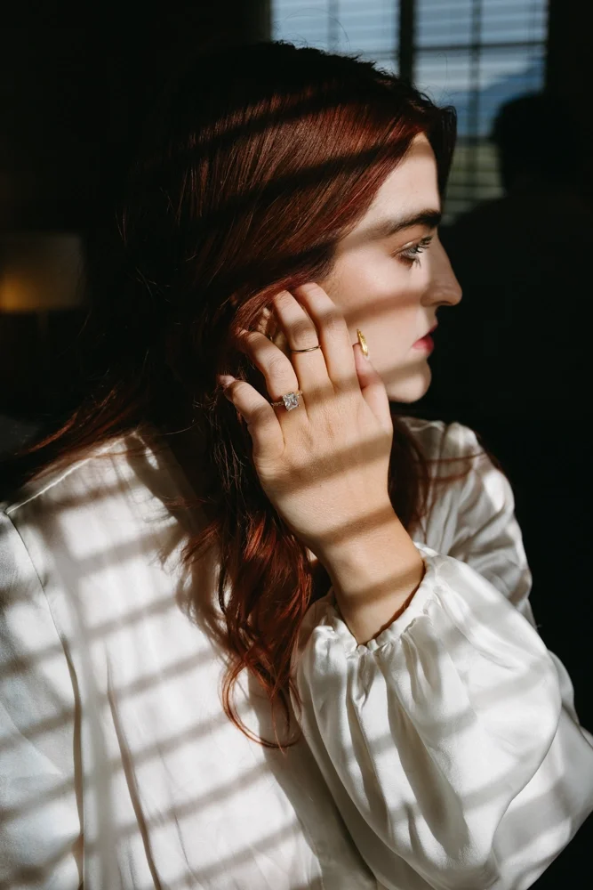 A woman with auburn hair and fair skin is sitting indoors by a window with blinds, casting striped shadows on her face. She is wearing a white blouse and is looking to the right, partially obscured by shadows.